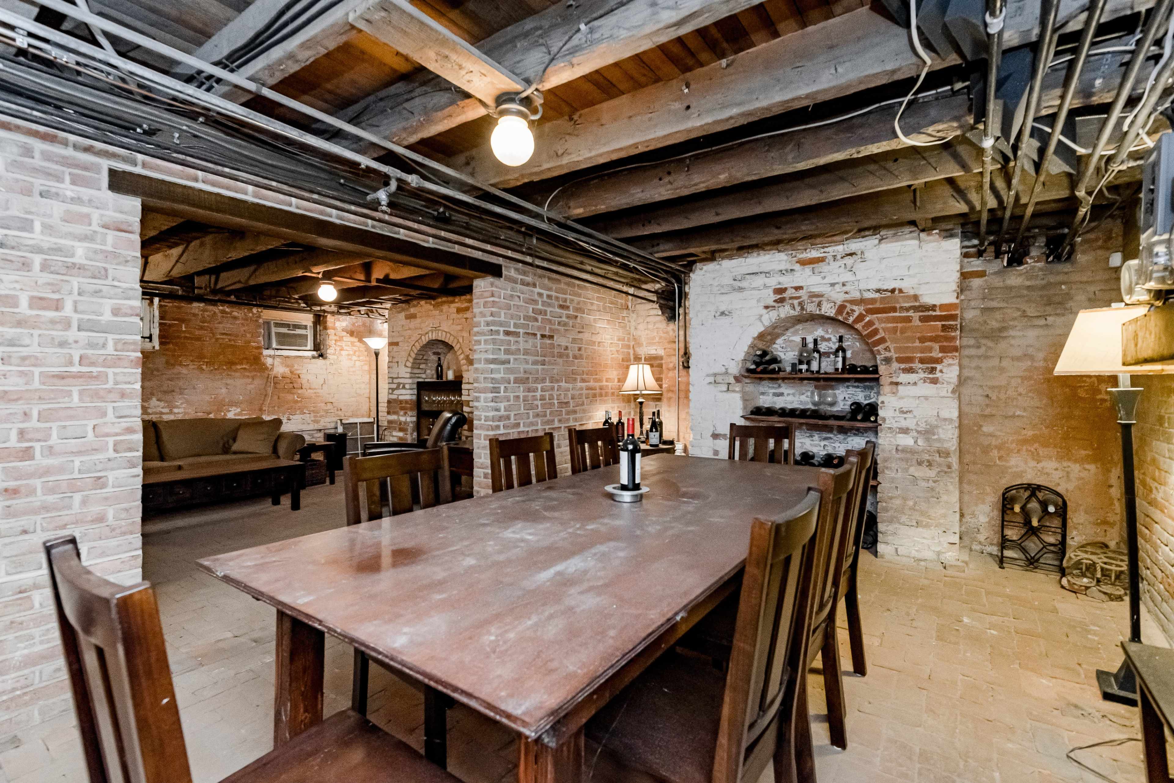 The image shows a spacious basement area featuring a wooden dining table surrounded by chairs, exposed brick walls, and various furnishings, including a lamp and shelves with bottles.