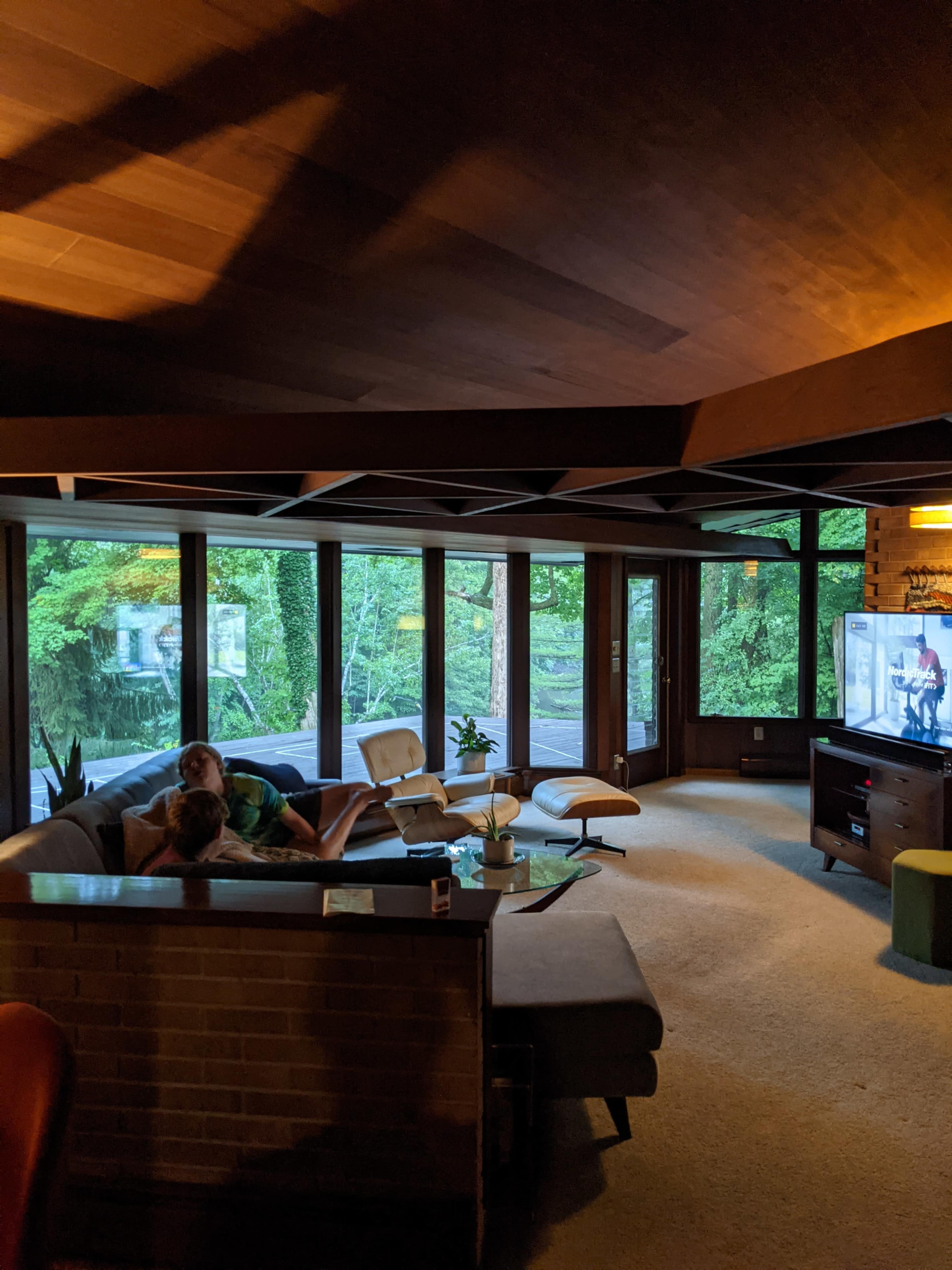 A cozy living room with large windows showcasing a view of greenery, featuring a sectional sofa, a television, and a coffee table.