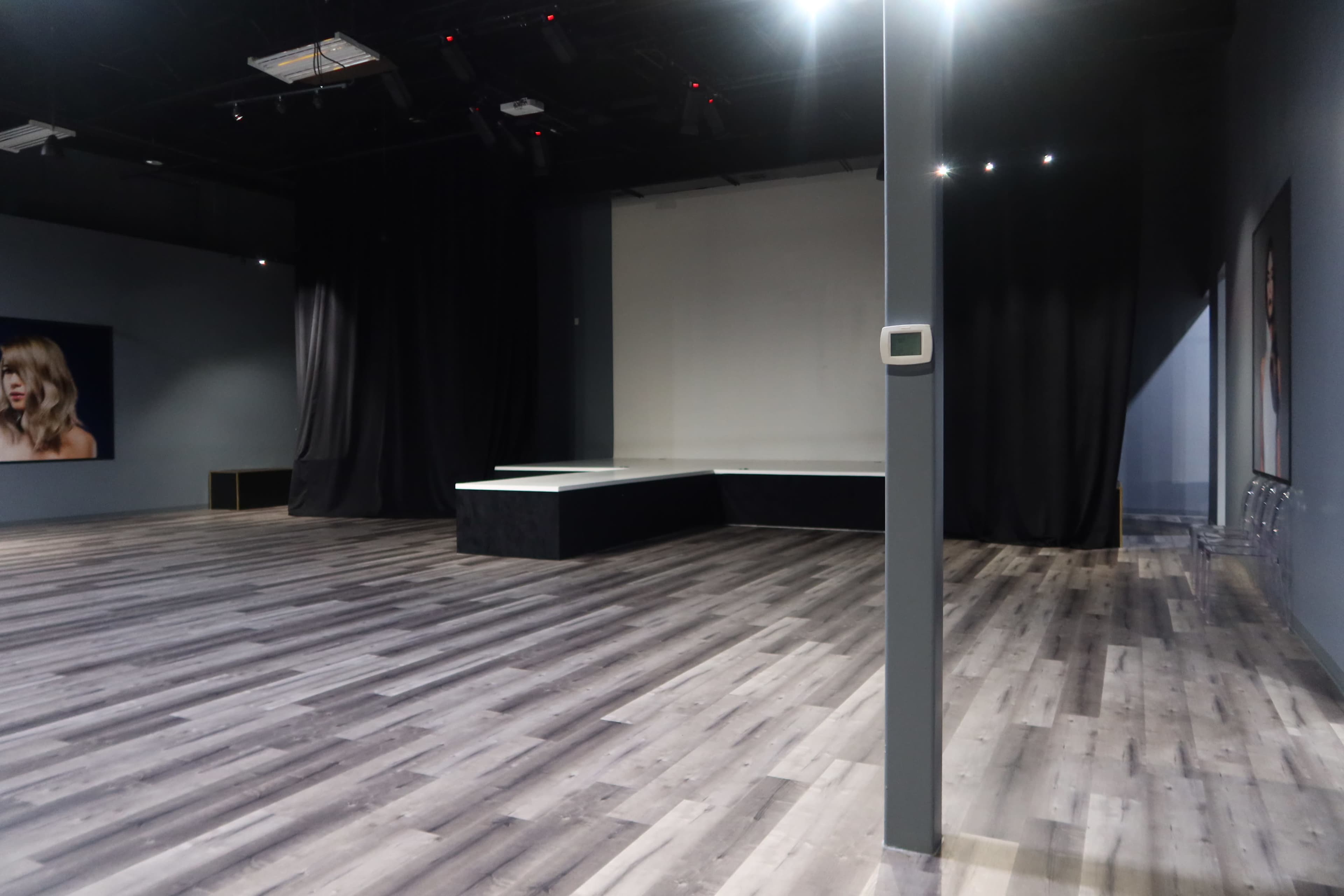 The image shows a spacious empty room with a wooden floor, featuring a simple stage at one end and dark curtains along the sides.