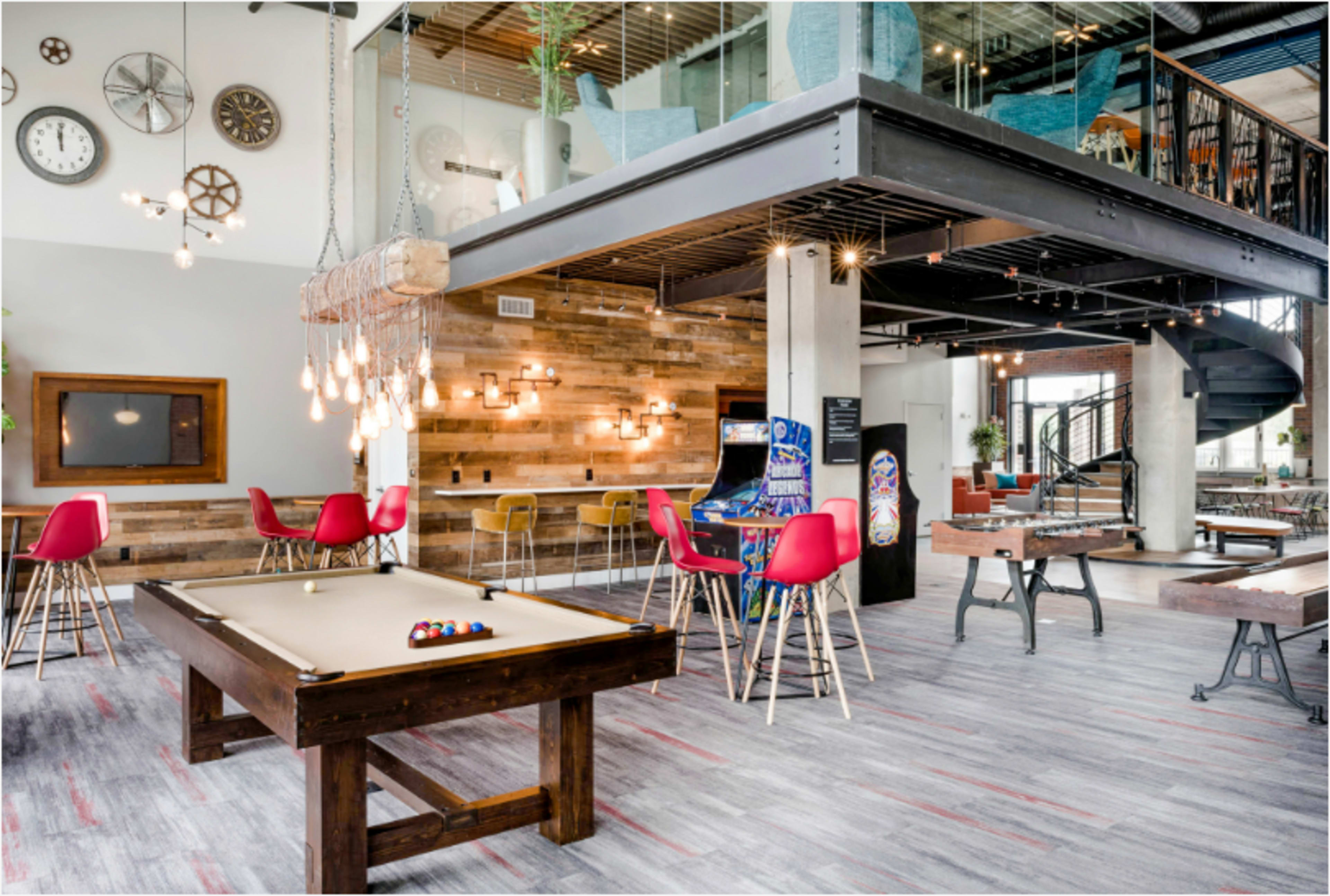 The image shows a modern recreational lounge area featuring a pool table, arcade games, and a mix of seating options, all within a beautifully designed space with industrial decor.
