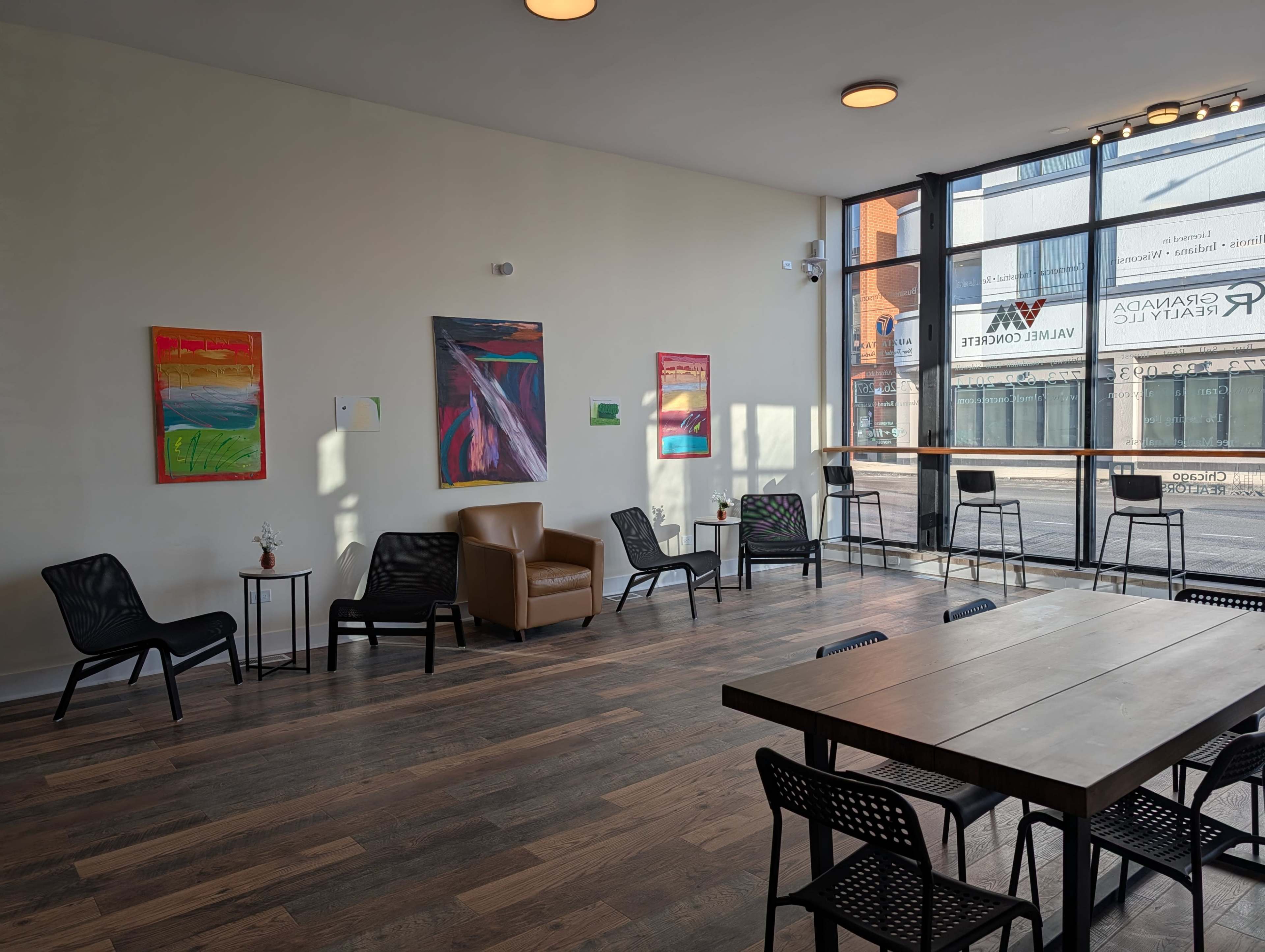 The image shows a bright, modern interior space with wooden flooring, several black chairs, a brown armchair, and colorful paintings on the walls near large windows.