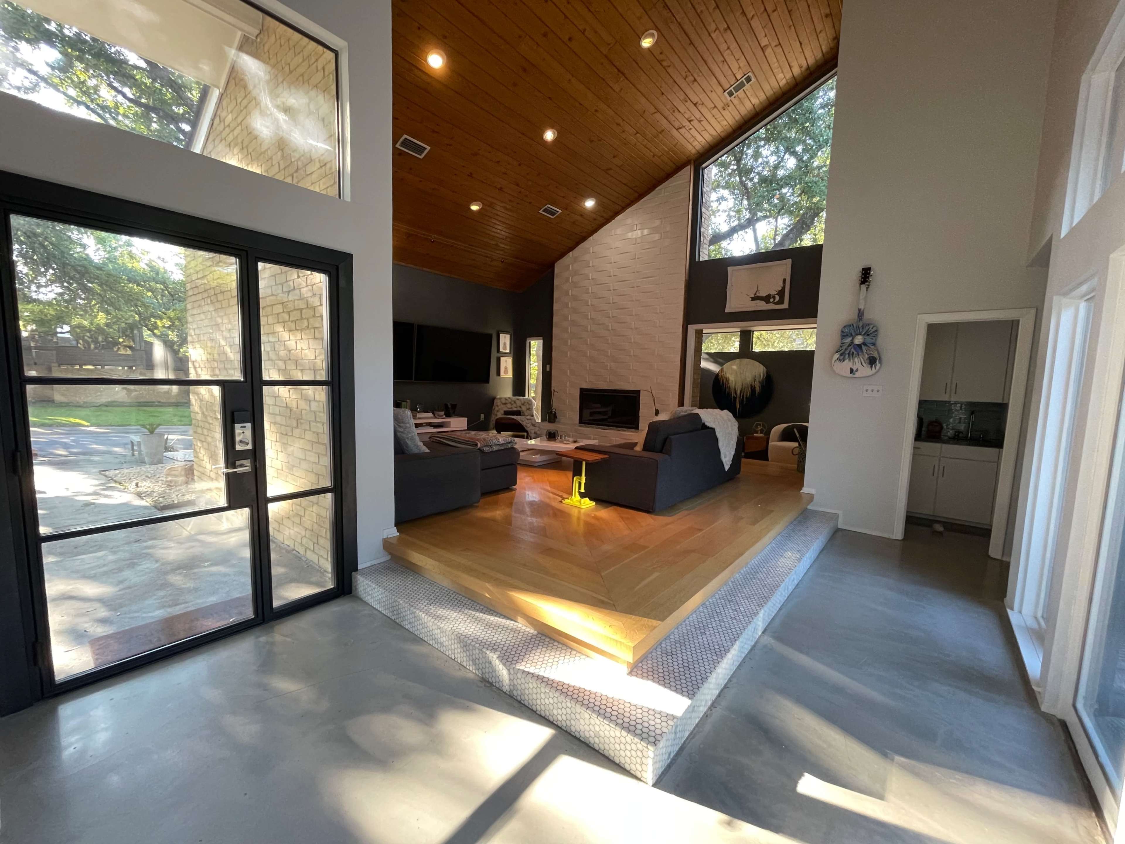 A contemporary living area with a high ceiling, a wooden beam structure, and a sunlit space featuring a fireplace and a transitional step leading to a kitchen area.