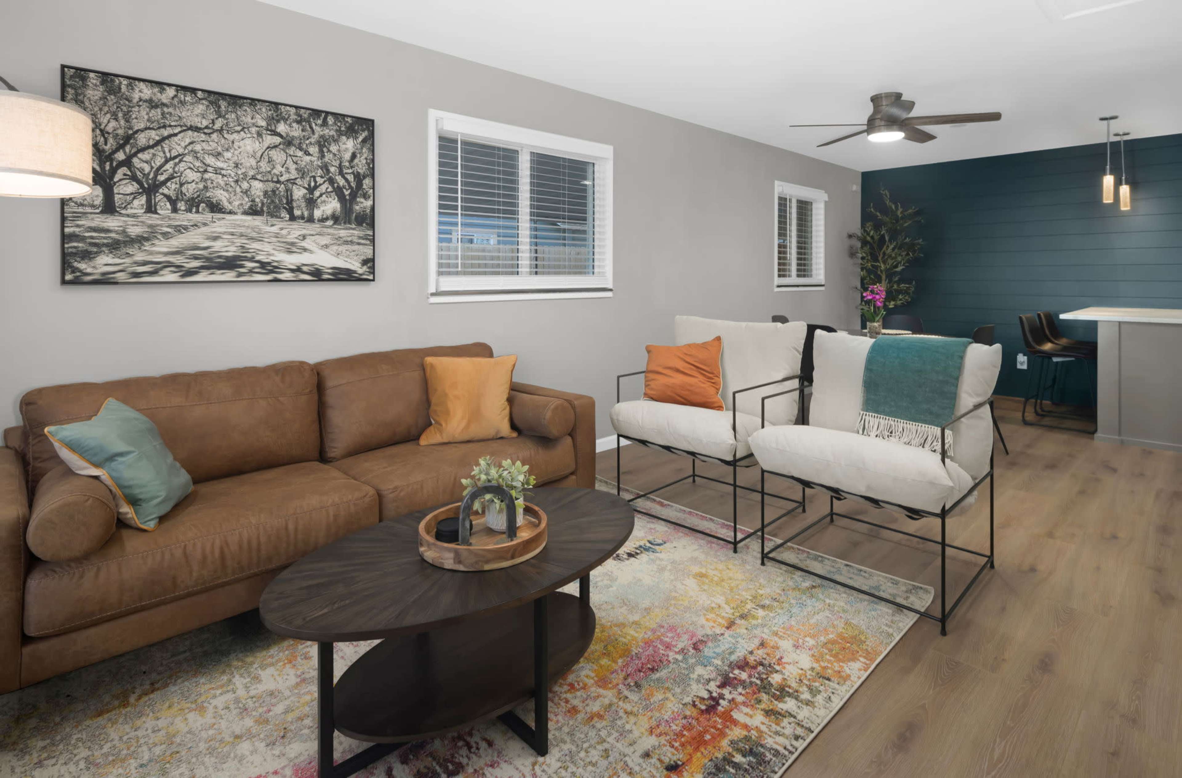 The image shows a modern living room with a brown leather sofa, two white chairs, a circular coffee table, and a colorful area rug, along with a dining area in the background.