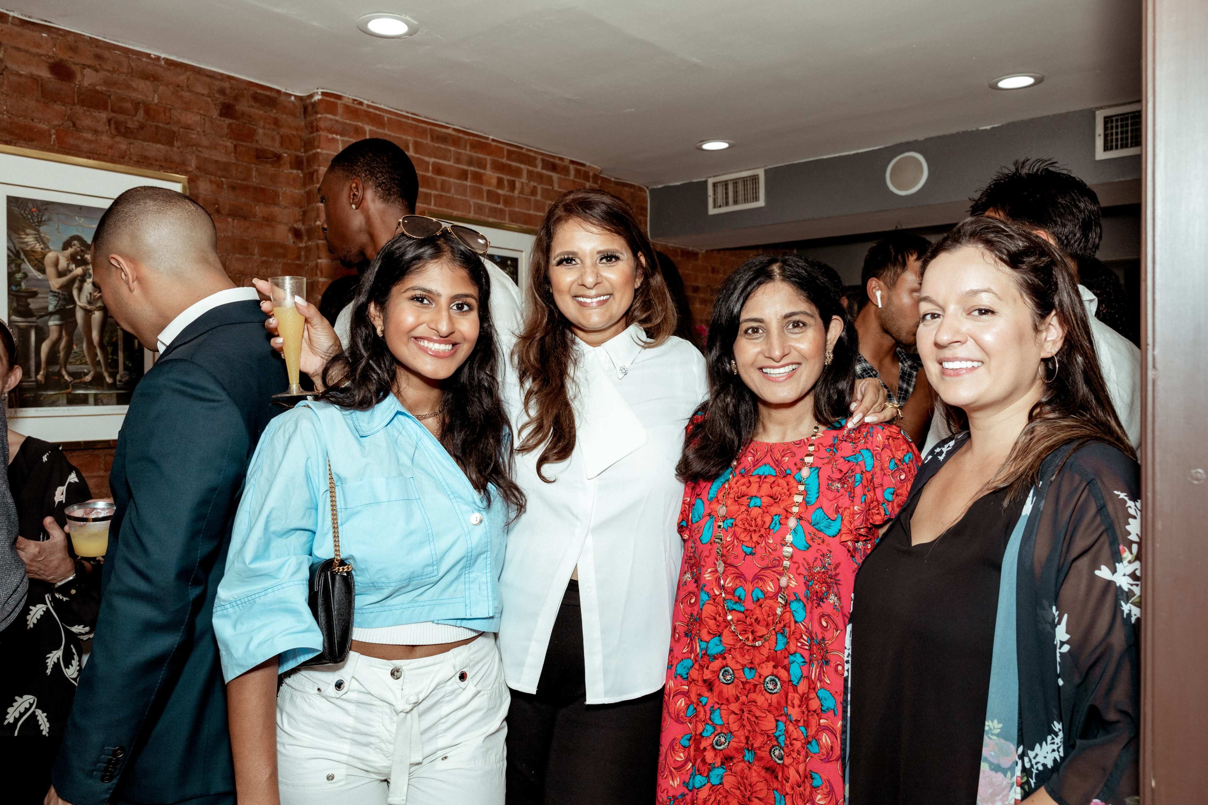 A group of four women poses together in a social setting with people mingling in the background.