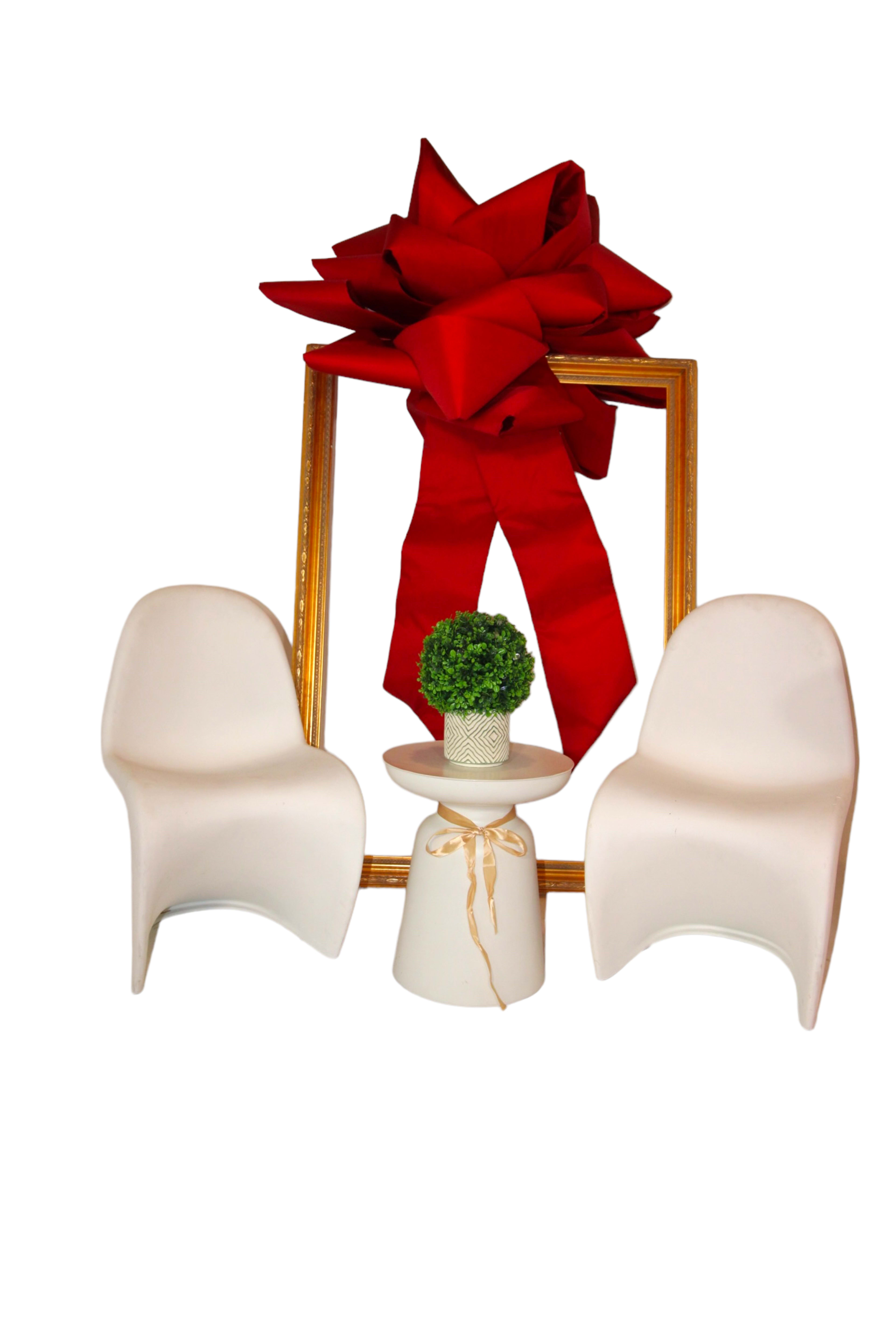 Two white chairs are positioned on either side of a small side table with a potted plant, framed by a large red bow above a gold frame.