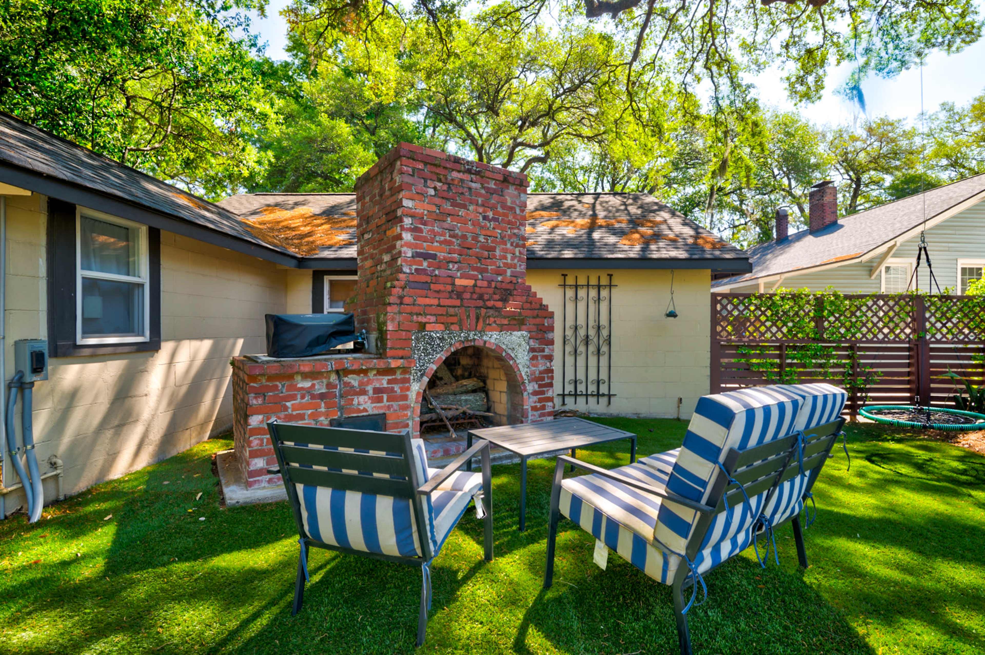 A brick outdoor fireplace sits in a grassy backyard, accompanied by two striped chairs and a small table.