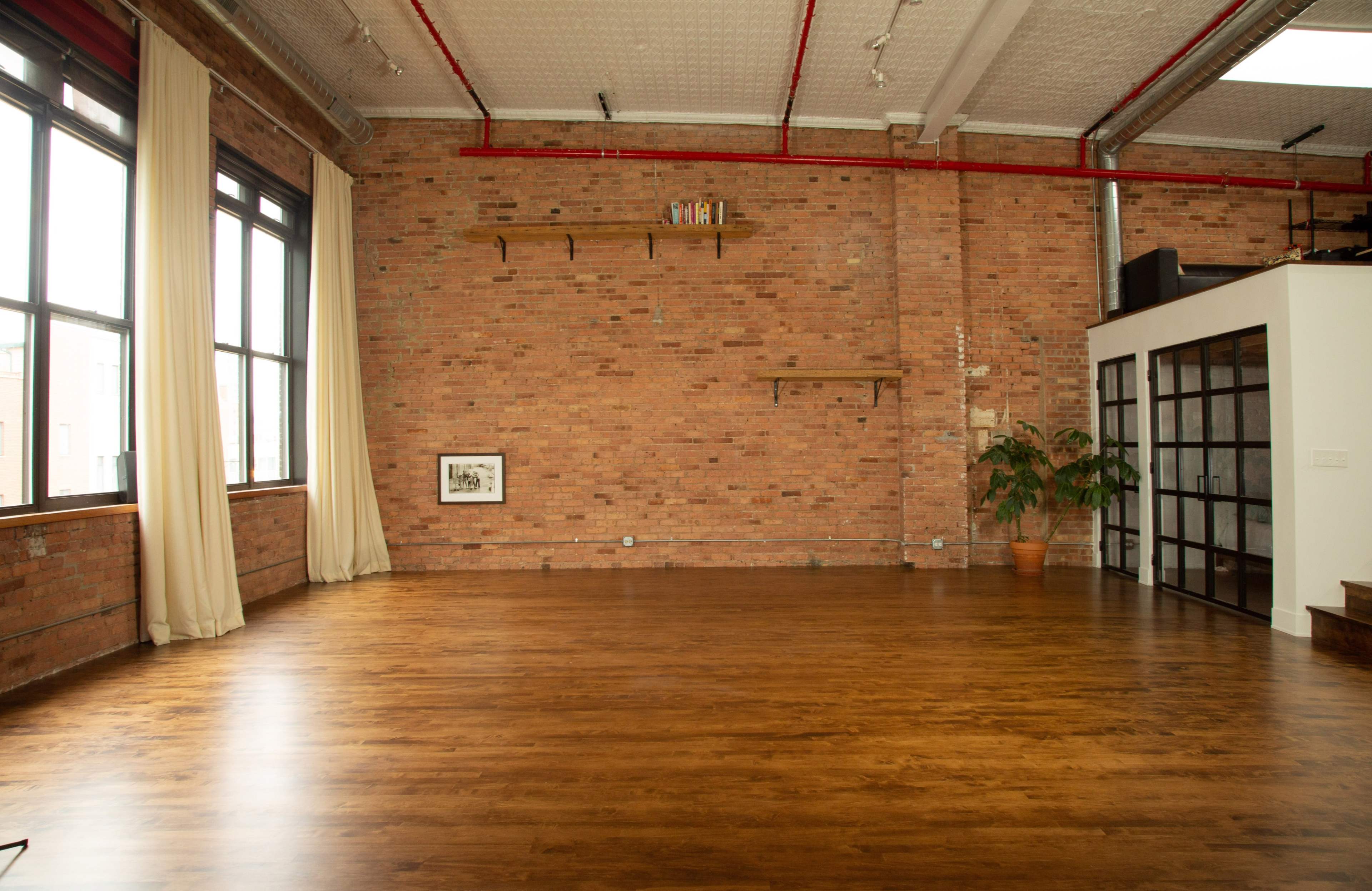 The image shows a spacious room with wood flooring, exposed brick walls, large windows, and a few decorative elements, including a plant and shelving units.