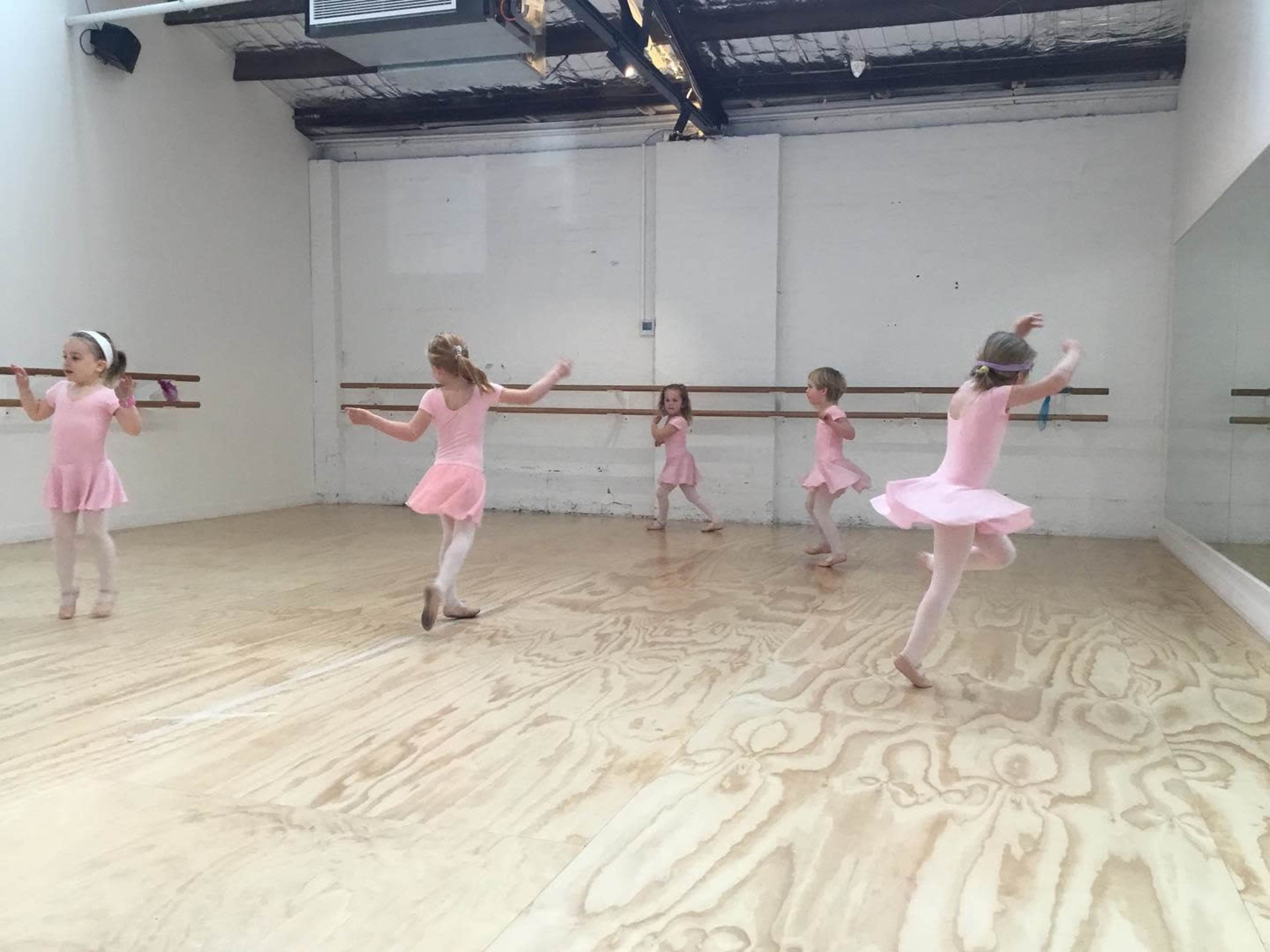 Five young girls in pink dance costumes are practicing ballet in a mirrored studio with wooden flooring.