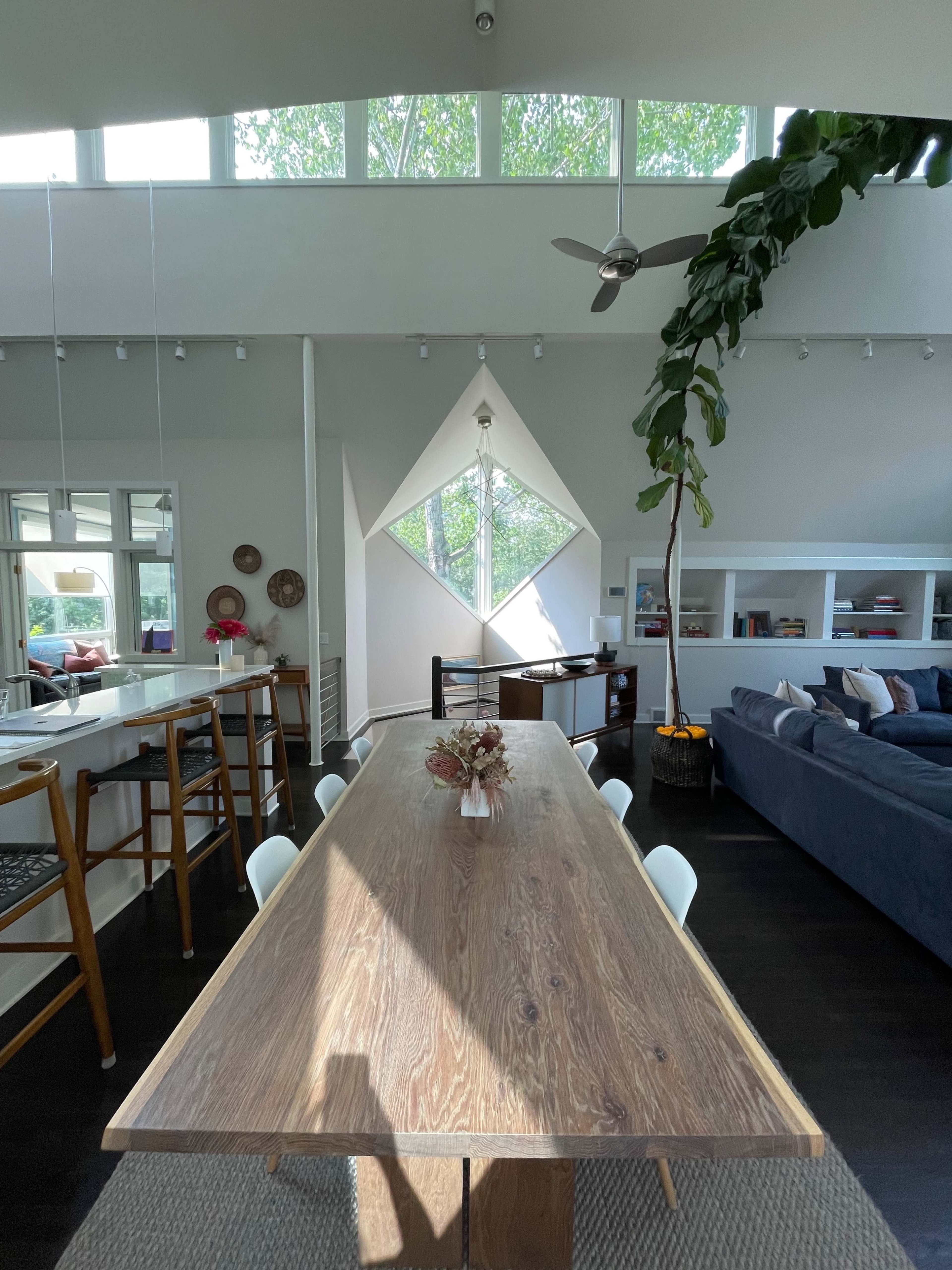 A modern interior features a long wooden dining table, a large diamond-shaped window, and a living area with a blue sofa and greenery.