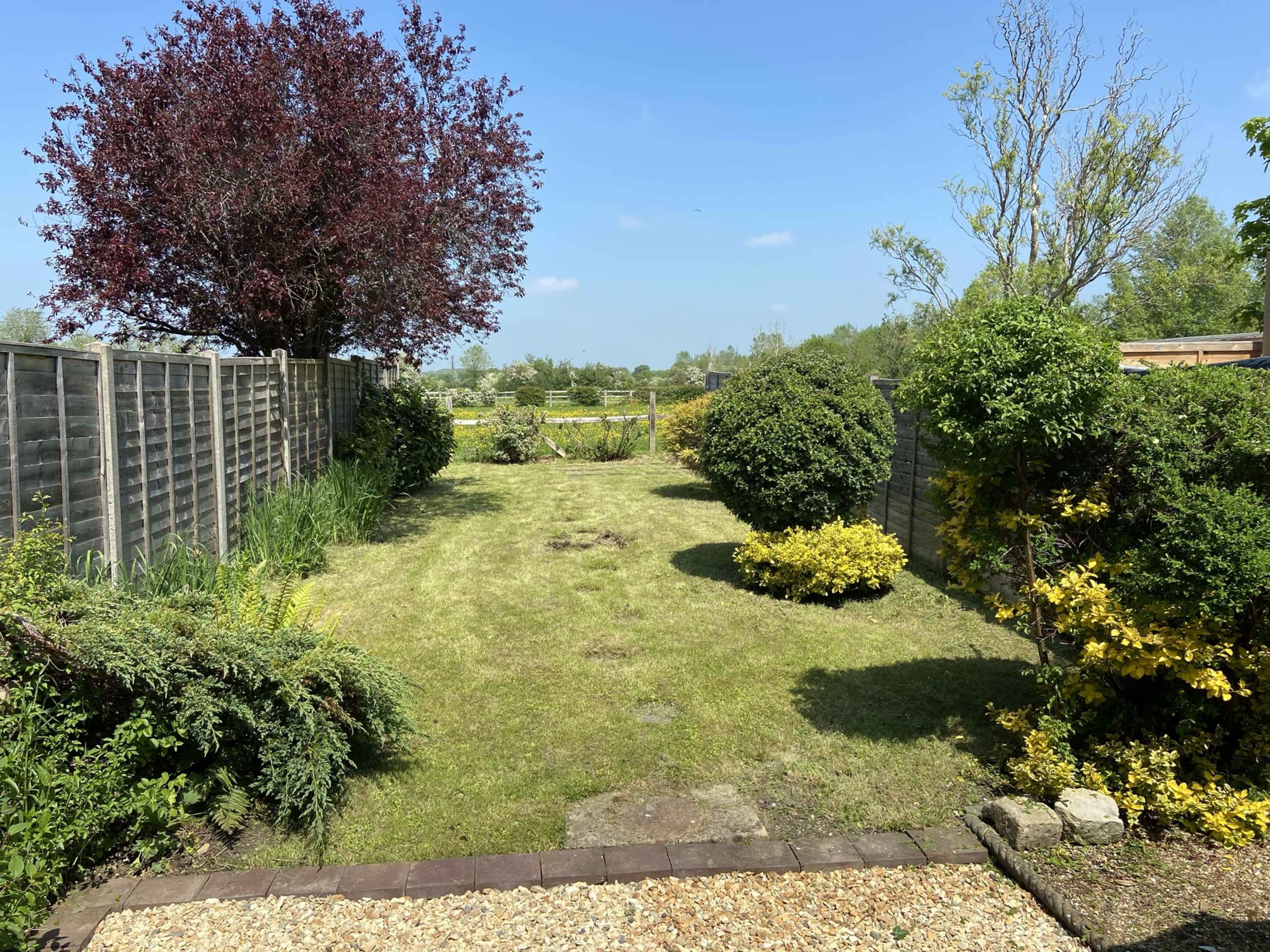 A well-maintained garden features a grassy path bordered by shrubs and trees, with a clear sky above.