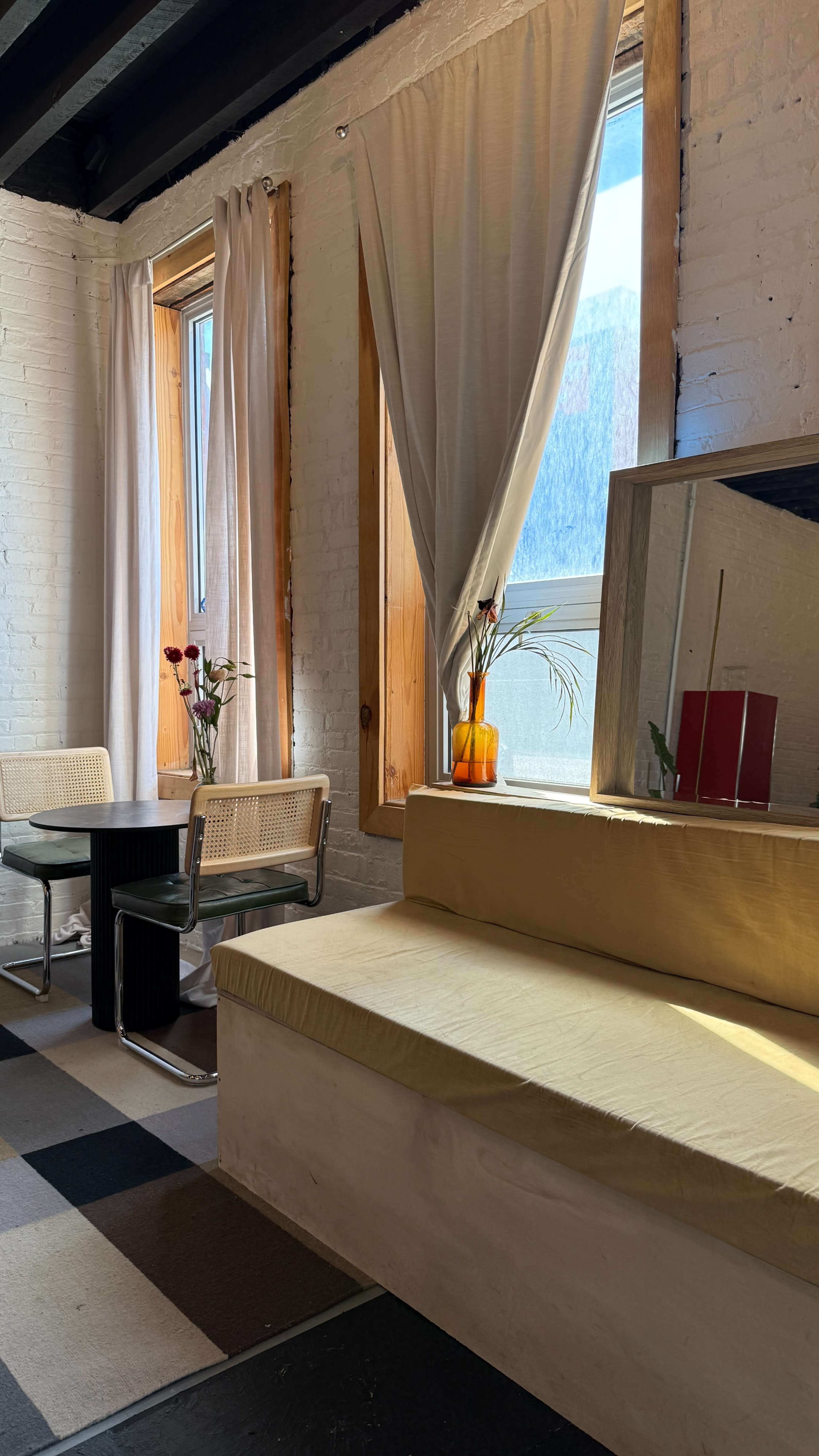 The image shows a bright room featuring two windows with white curtains, a small dining table with chairs, and a beige bench against a wall.