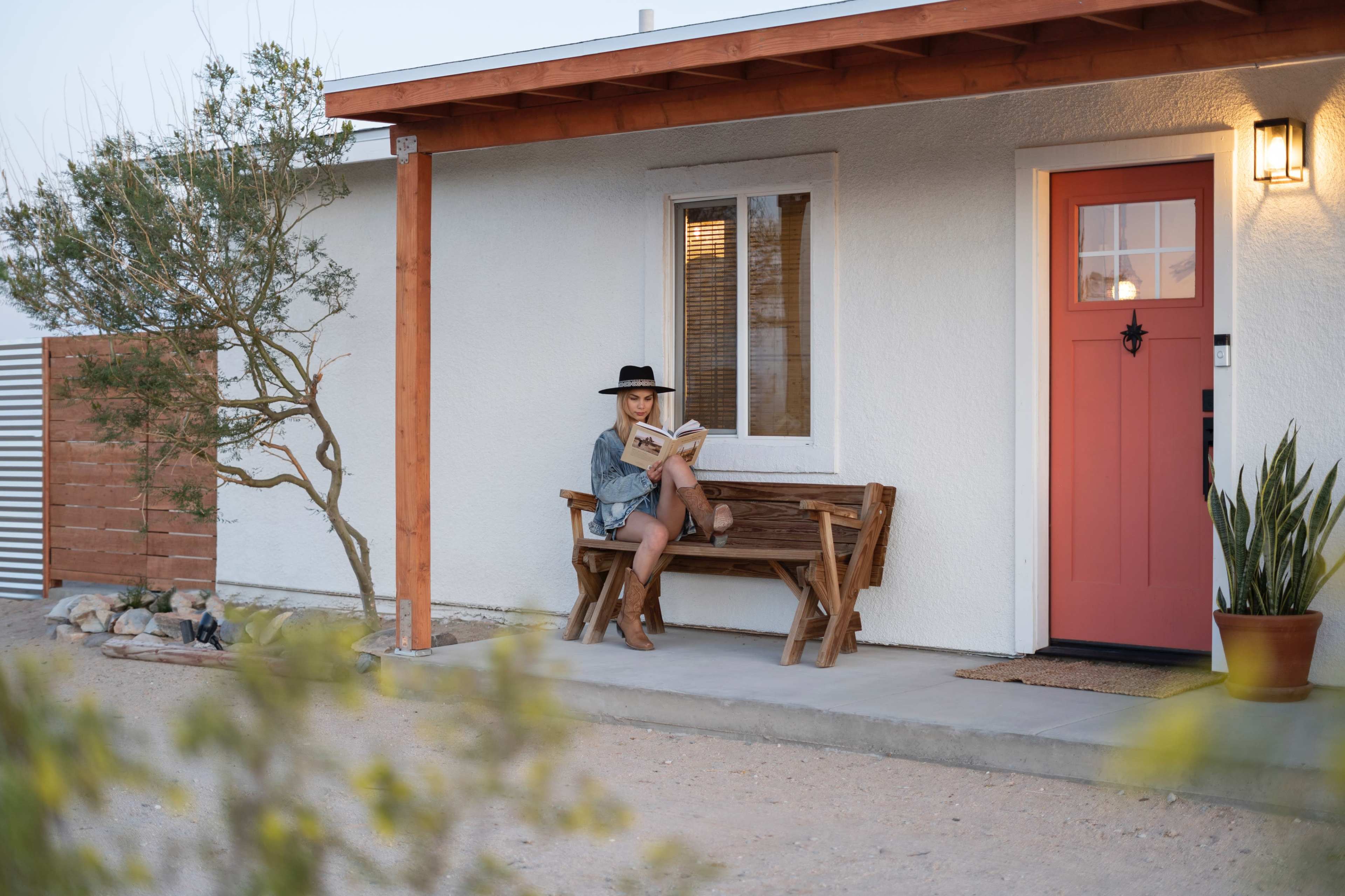A person sitting on a bench outside a house, reading a book, with a hat on and a potted plant nearby.