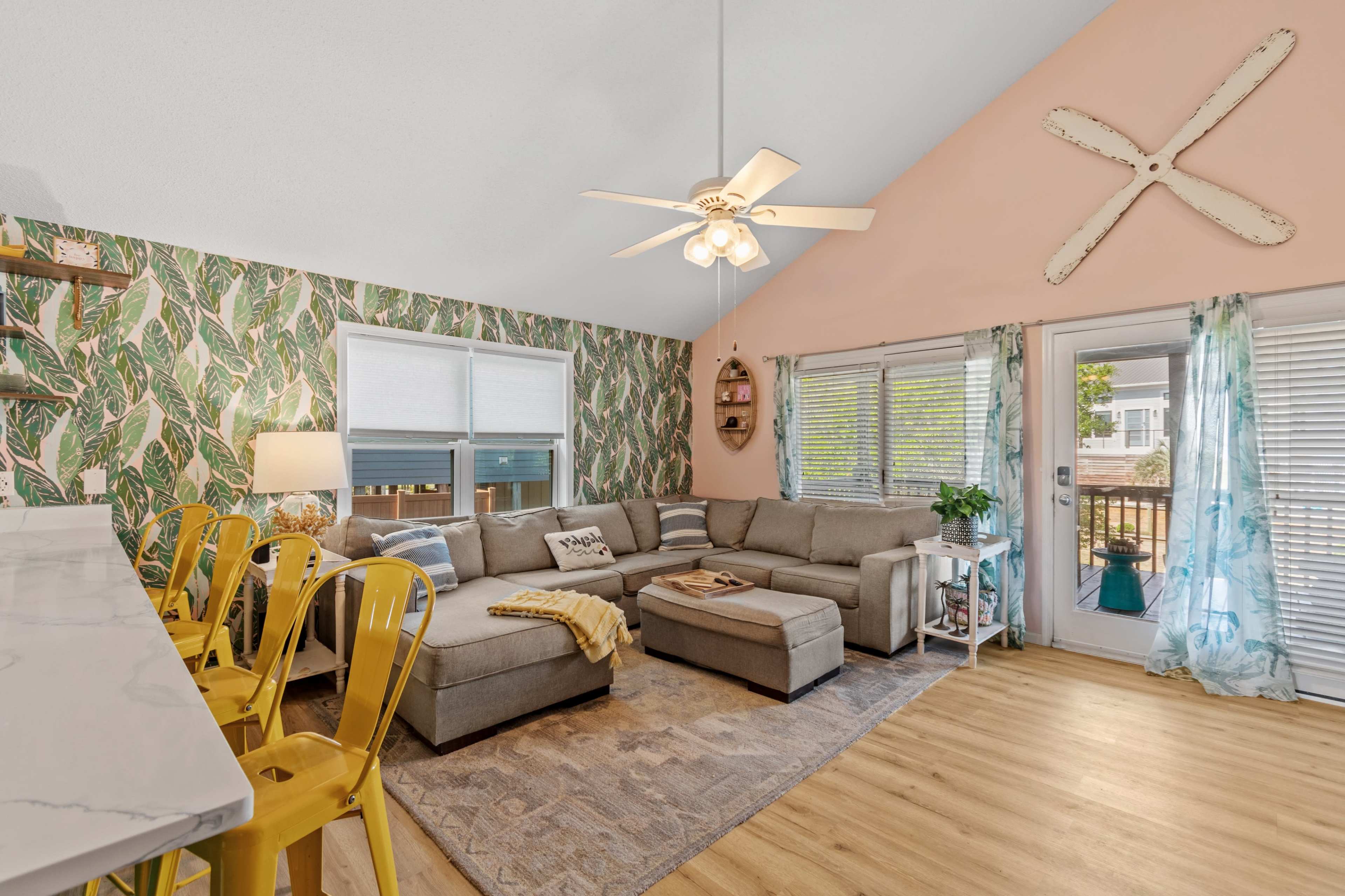 The image shows a bright living room with a sectional sofa, a dining area featuring yellow chairs, and large windows that provide natural light, all accented by tropical-themed wallpaper.
