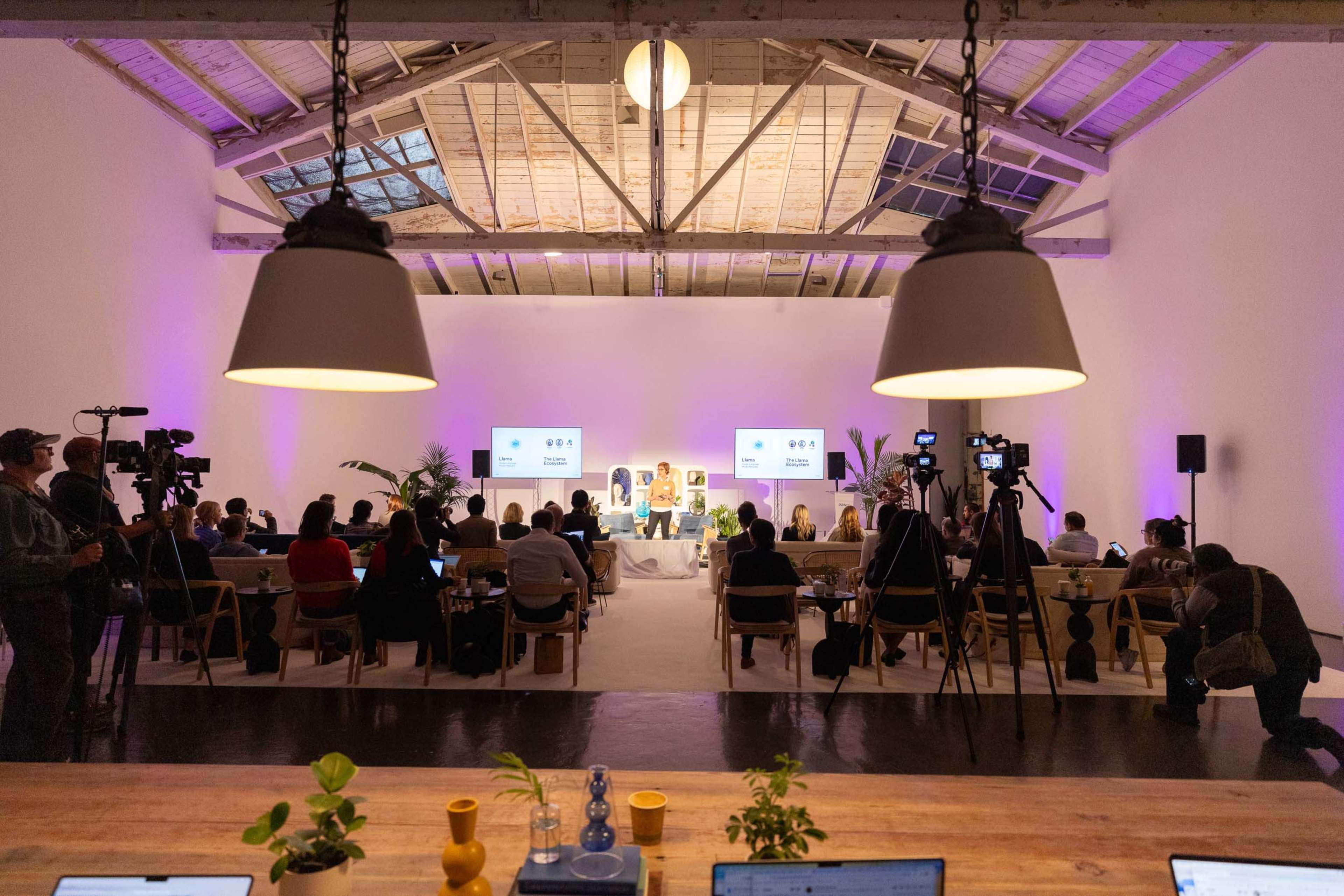 A conference event is taking place in a spacious venue with a panel on stage and an audience seated, illuminated by purple lighting.