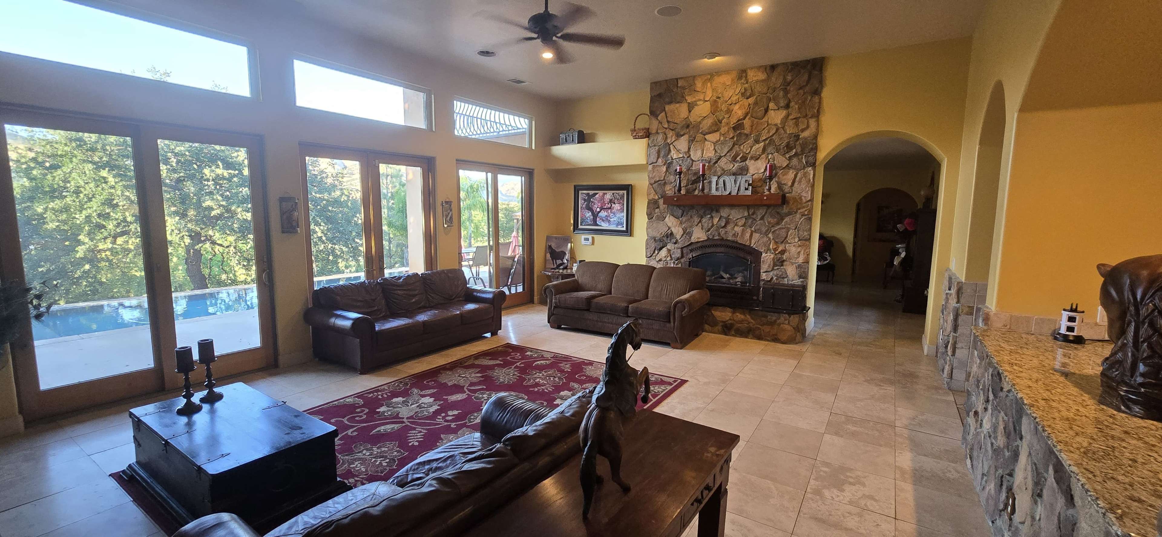 The spacious living room features large windows, a stone fireplace, and a combination of brown leather and fabric seating arranged around a decorative area rug.
