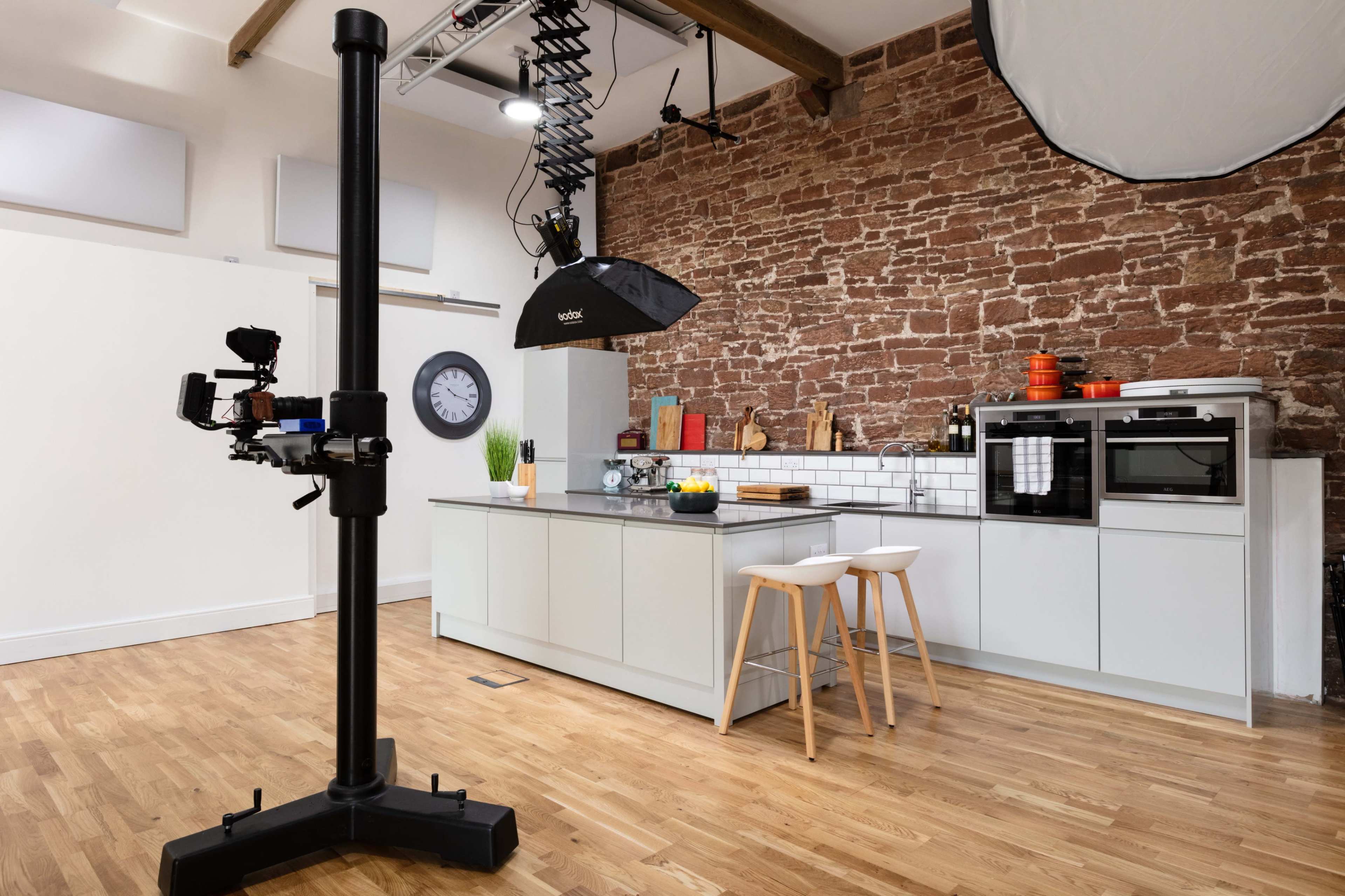 The image shows a modern kitchen with a brick wall, featuring white cabinetry, a central island with stools, and professional lighting equipment set up for a photoshoot.