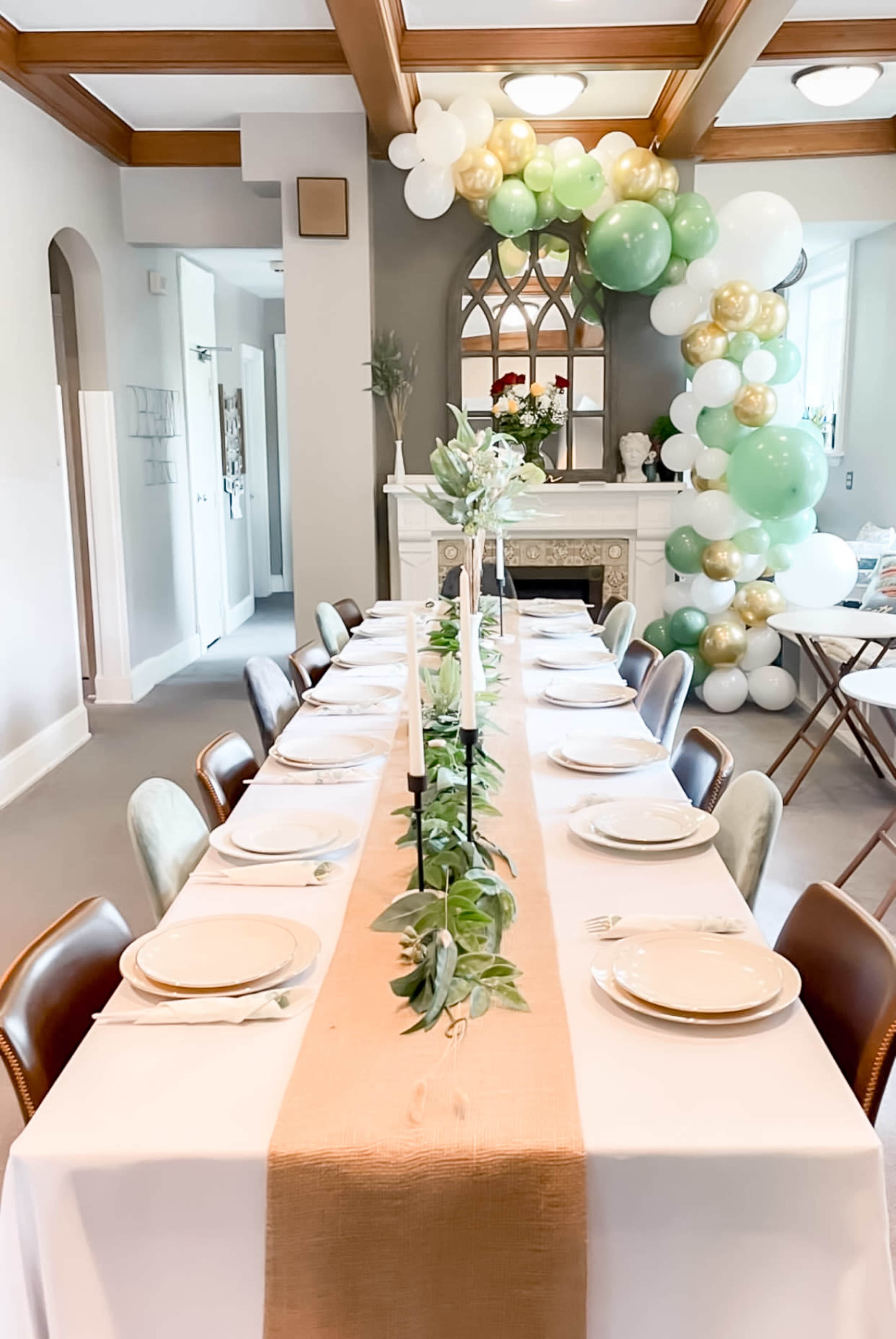 A long dining table is set with white tablecloths and place settings, featuring a centerpiece of greenery, while a balloon arch decorates the space behind a fireplace.