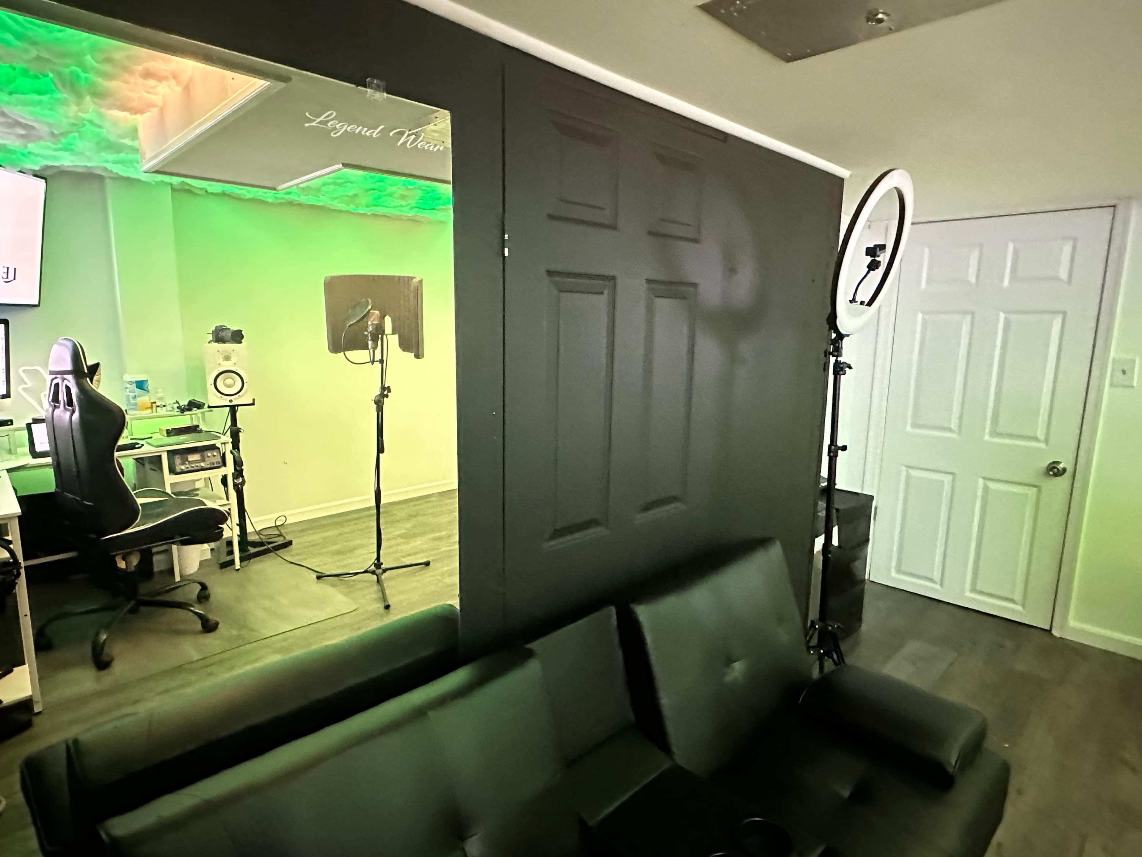 A room with a black wall, a large mirror reflecting a green-lit workspace, and a ring light next to a closed door.
