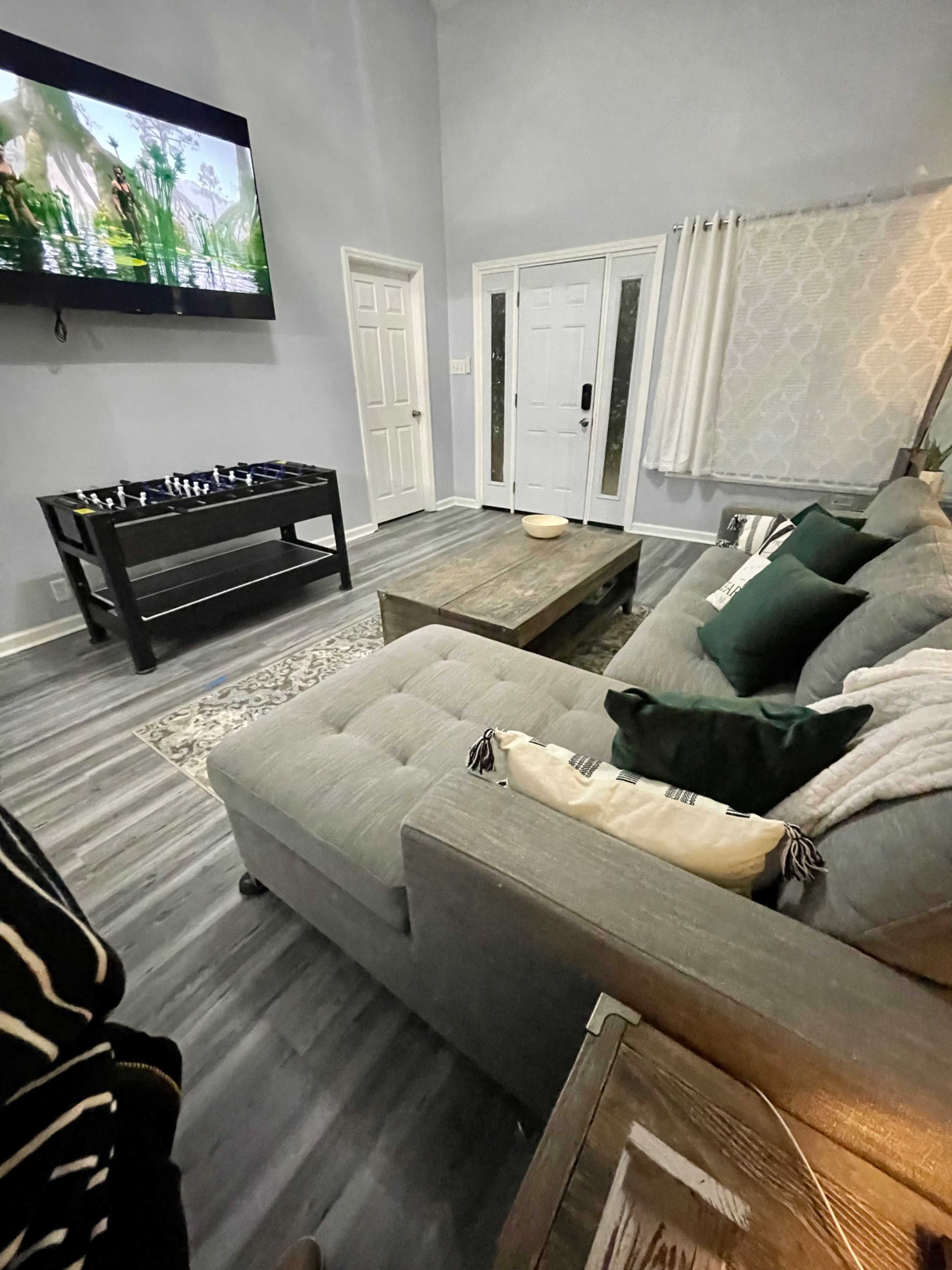 A living room featuring a grey sofa, a wooden coffee table, a television mounted on the wall, and a front door visible in the background.