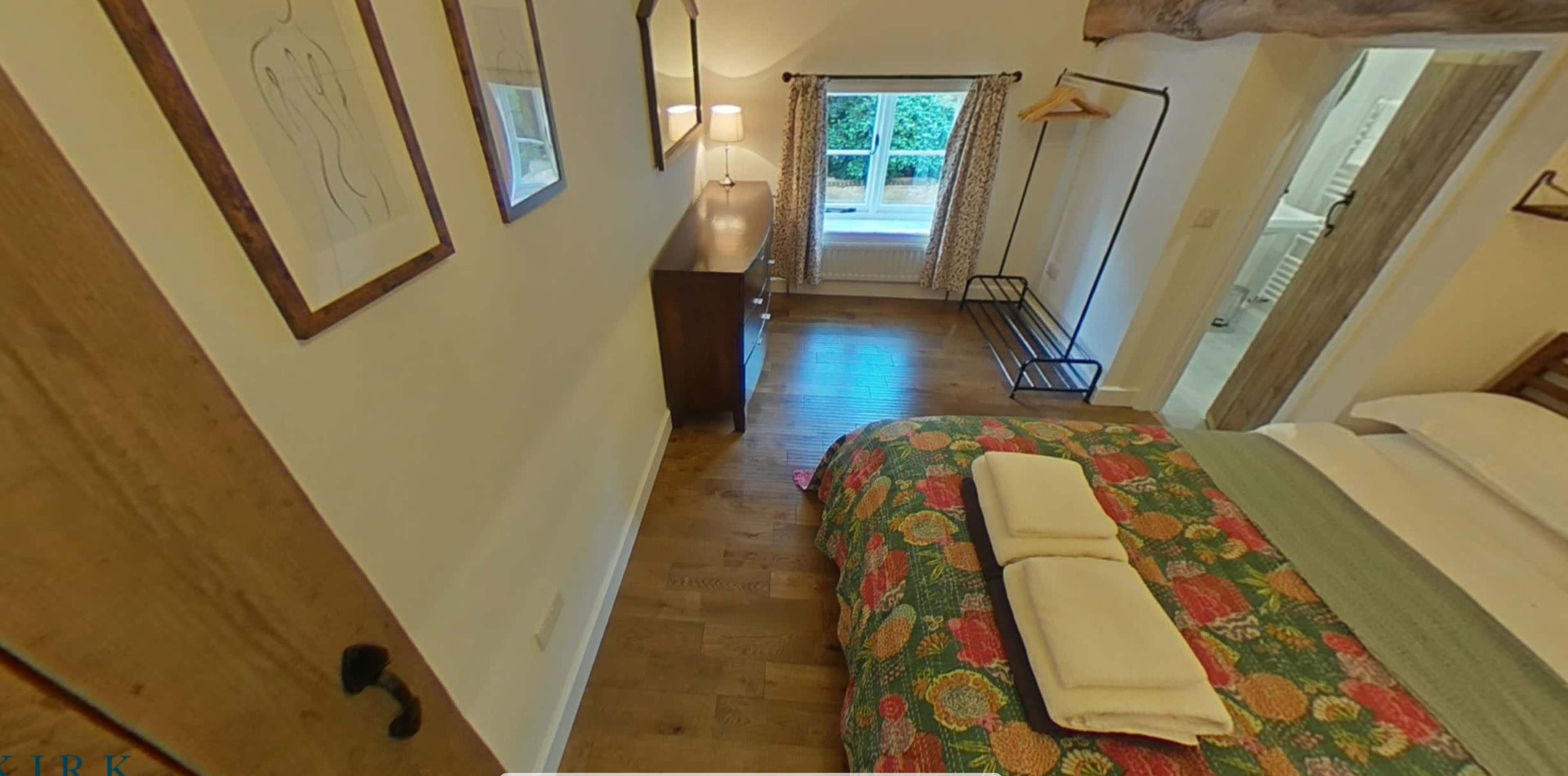 A bedroom with a colorful bedspread, a wooden dresser, and a window revealing greenery outside.