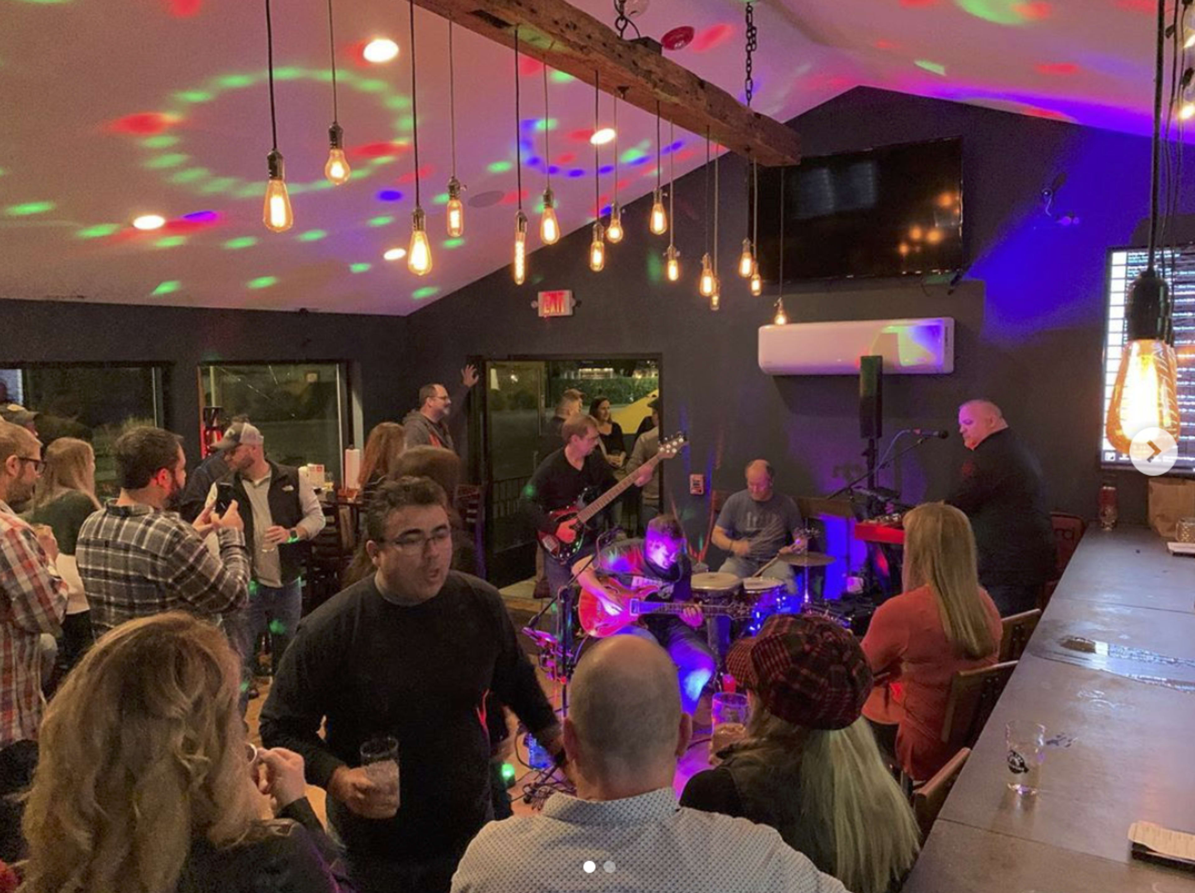 A lively band performs in a bar filled with an engaged audience, illuminated by colorful lights and hanging bulbs.