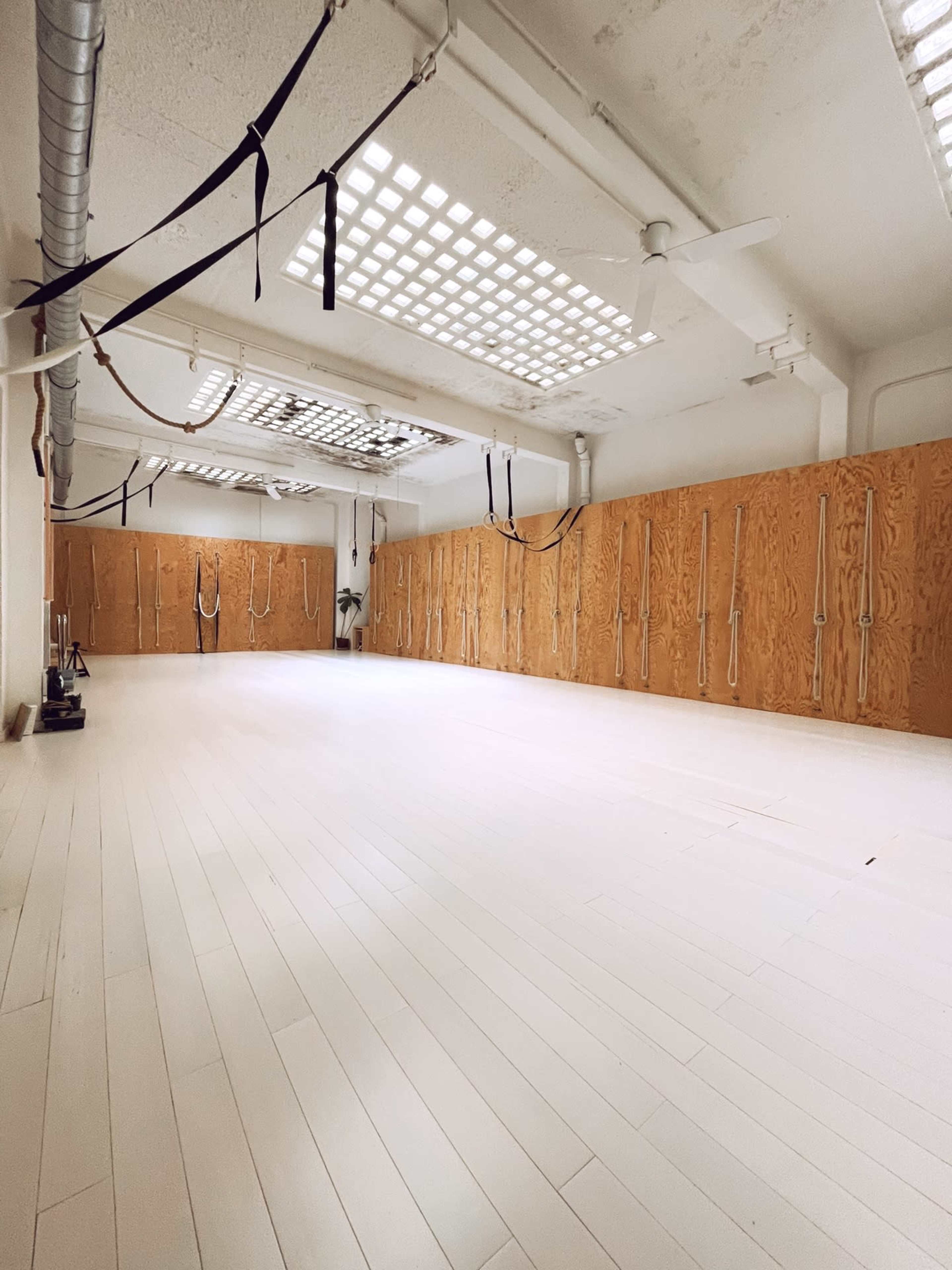 A spacious, empty yoga studio with white wooden floors and wooden wall panels featuring ropes hanging on hooks.