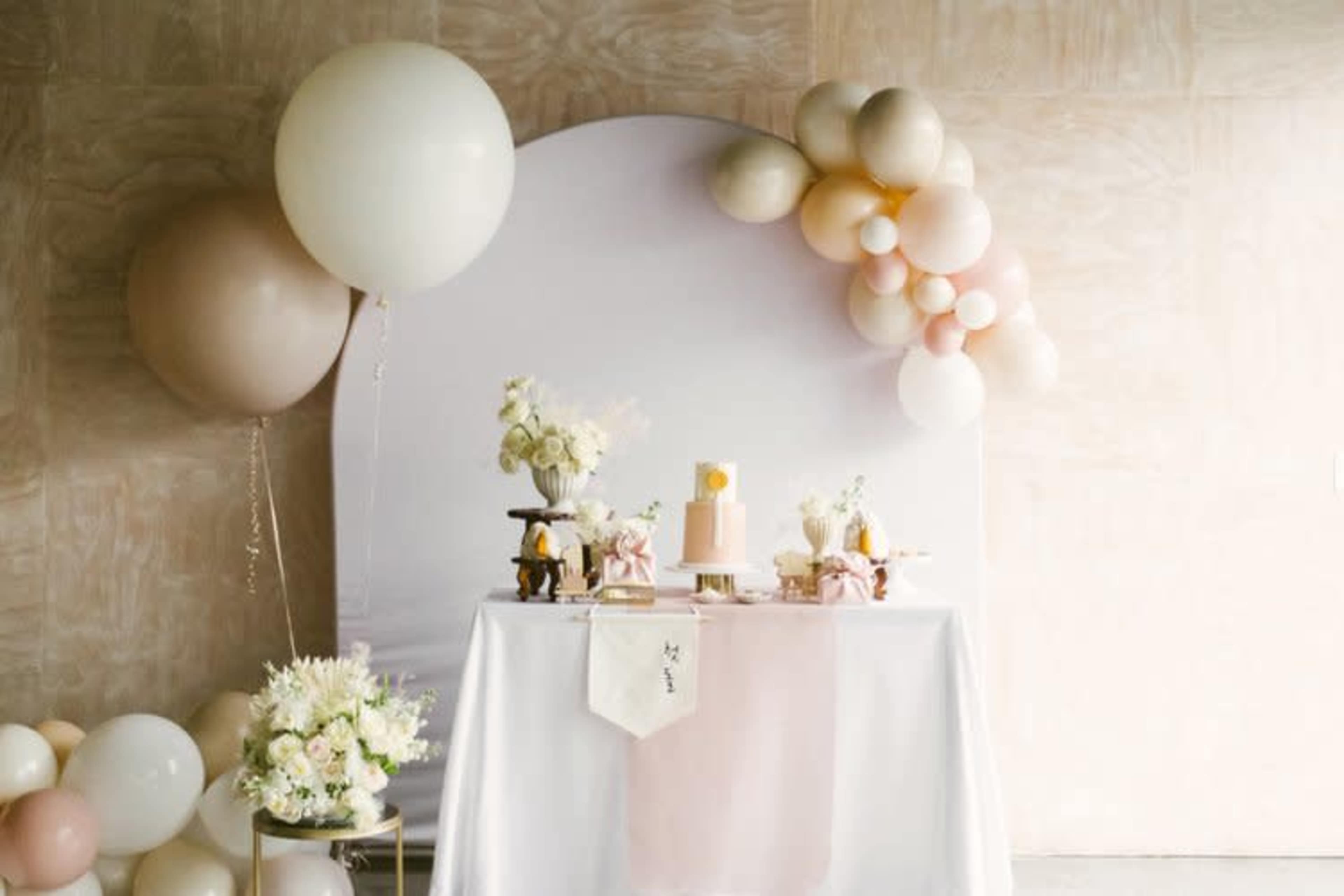 A decorative table is set up with a cake and floral arrangements, accompanied by pastel balloons and a backdrop.