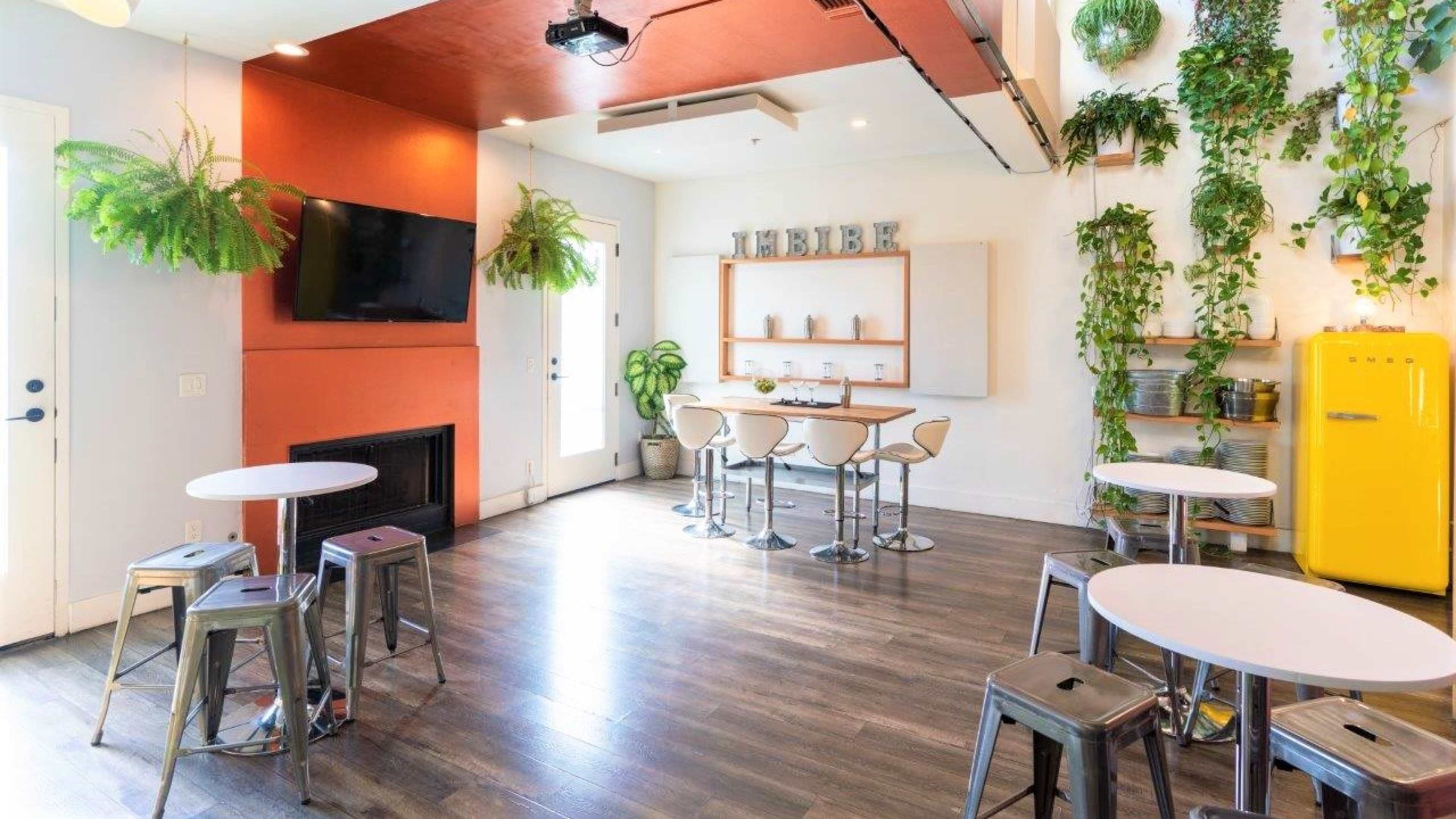 A bright room features a yellow refrigerator, white tables, metal stools, and several plants hanging from the ceiling.