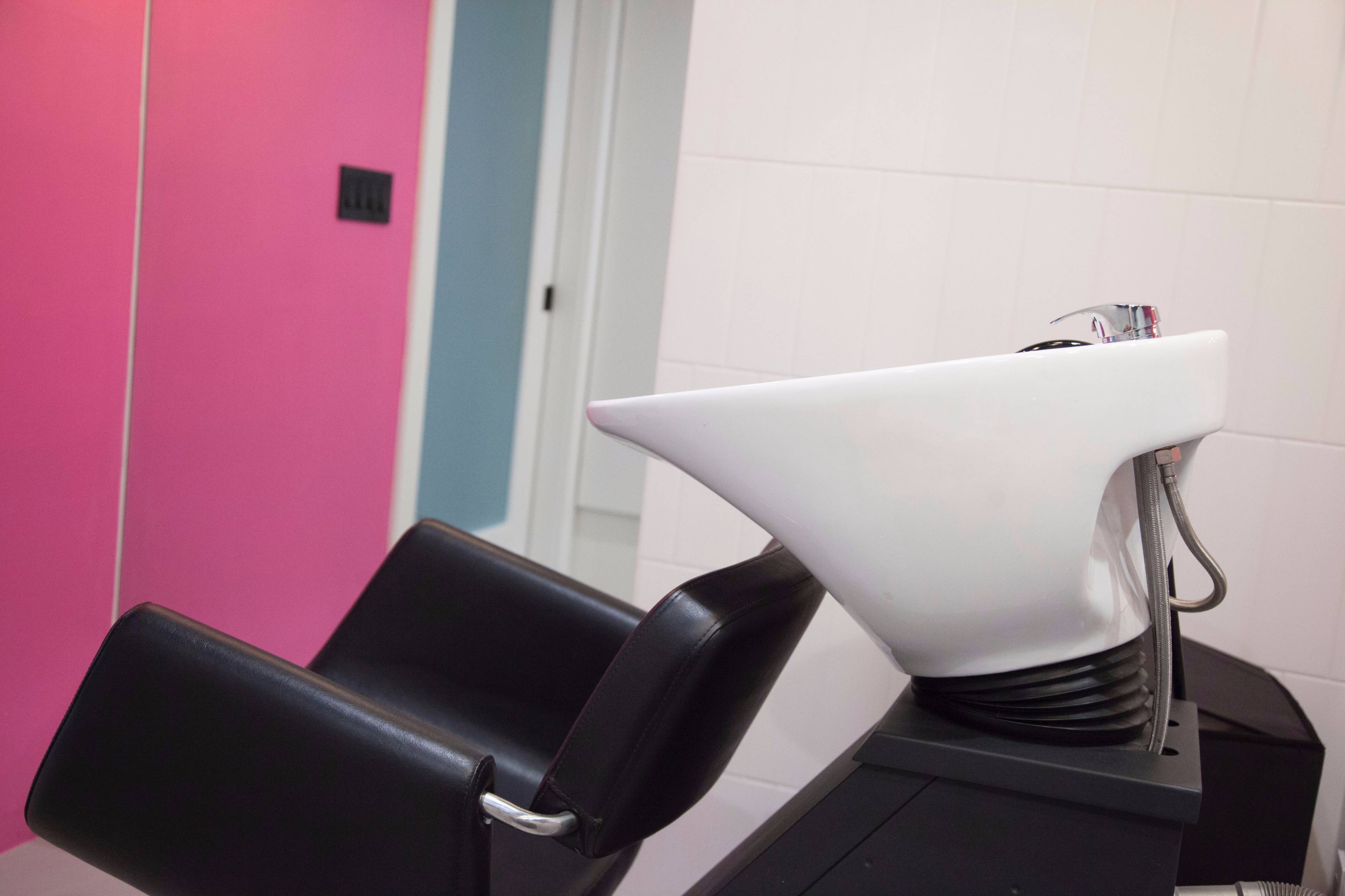 A black salon chair is positioned in front of a white shampoo basin against a wall with pink and blue accents.
