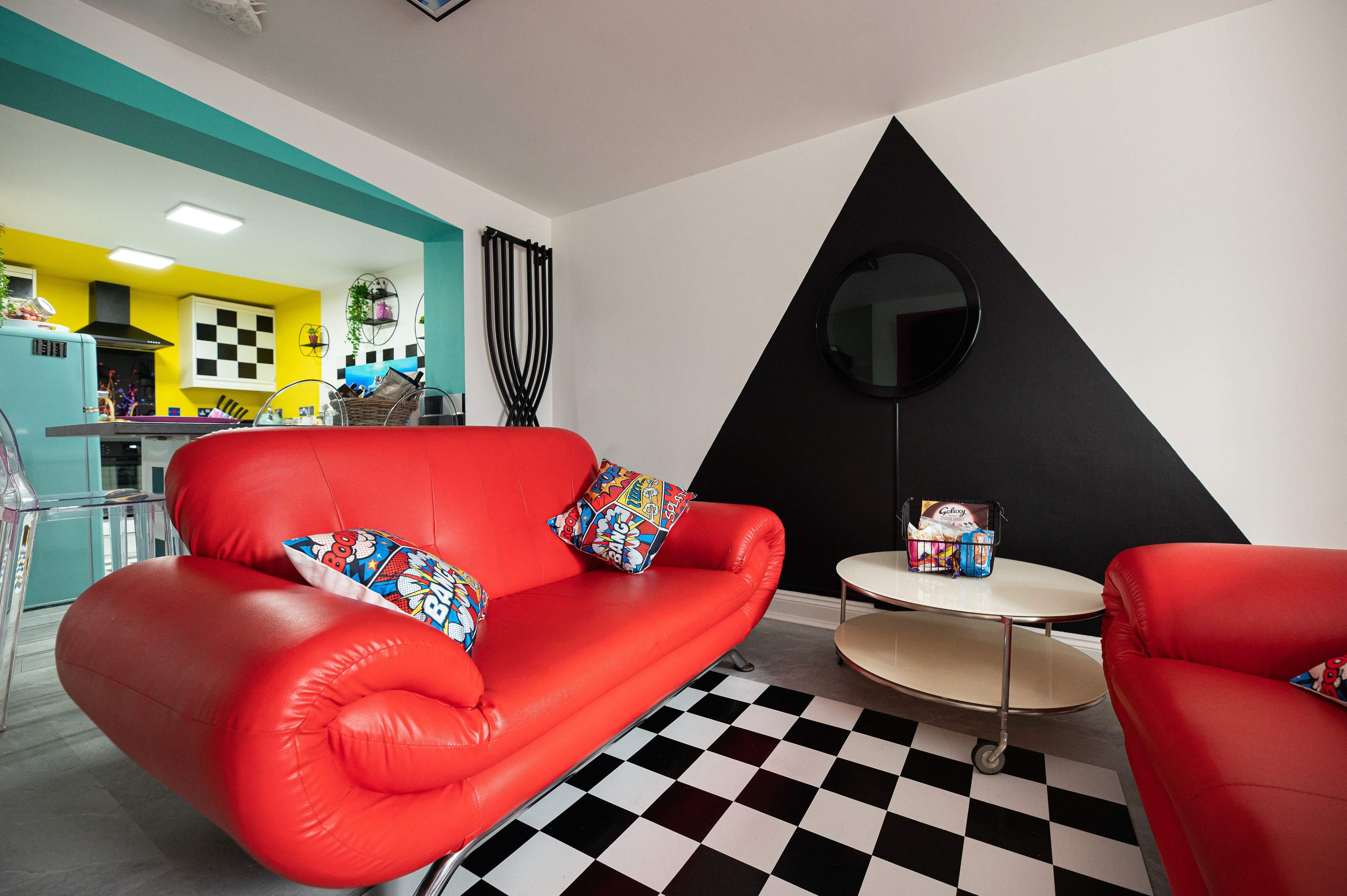 A brightly colored living space featuring red sofas with decorative pillows, a round coffee table, and a black and white checkered floor, set against a backdrop of yellow and black walls.