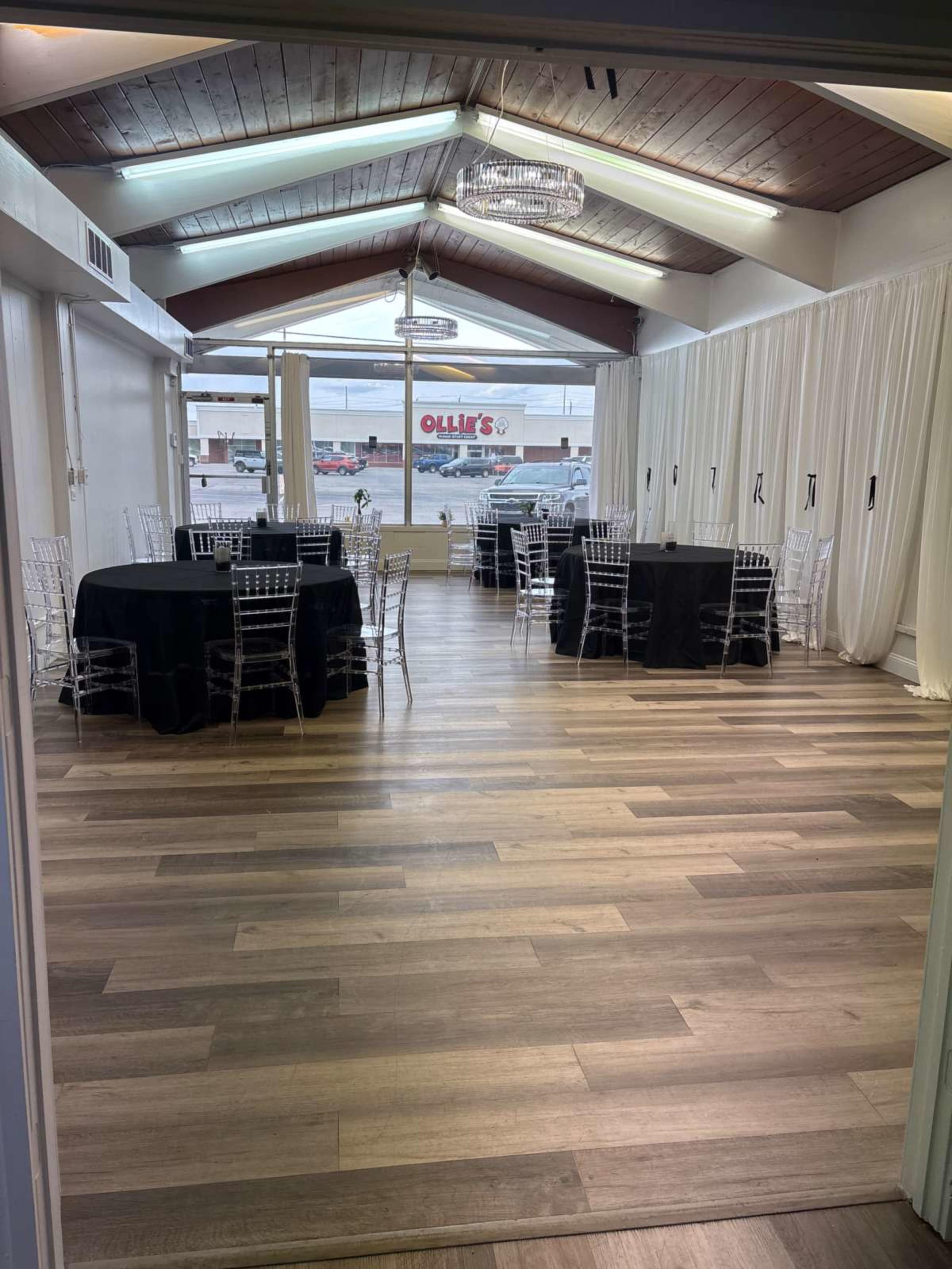 The image shows a spacious event room with black and clear chairs arranged around round tables, adorned with elegant table settings, and large windows allowing natural light to enter.