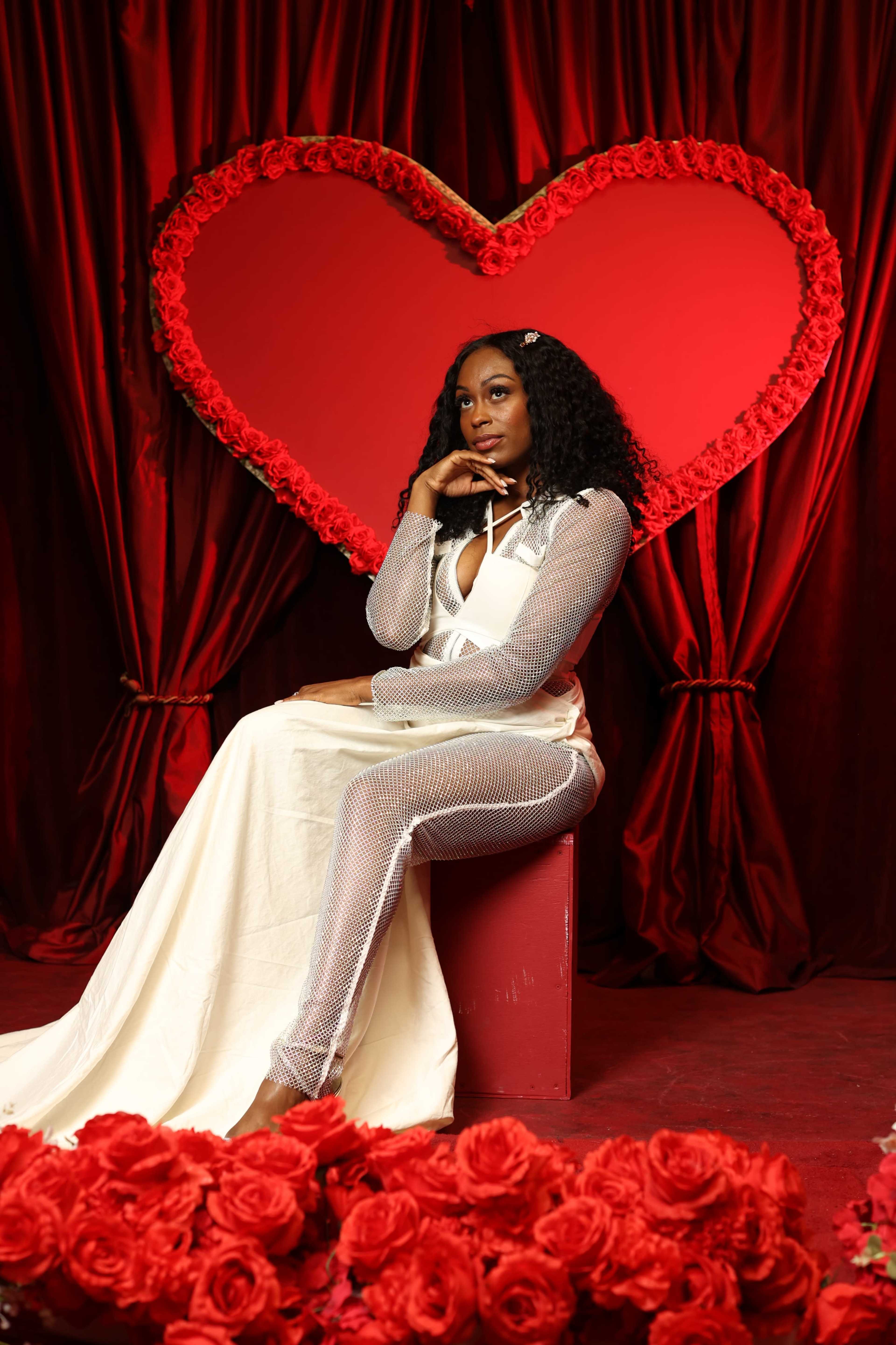 A woman in a form-fitting outfit sits on a red box in front of a large heart made of roses, surrounded by rose petals on the ground.