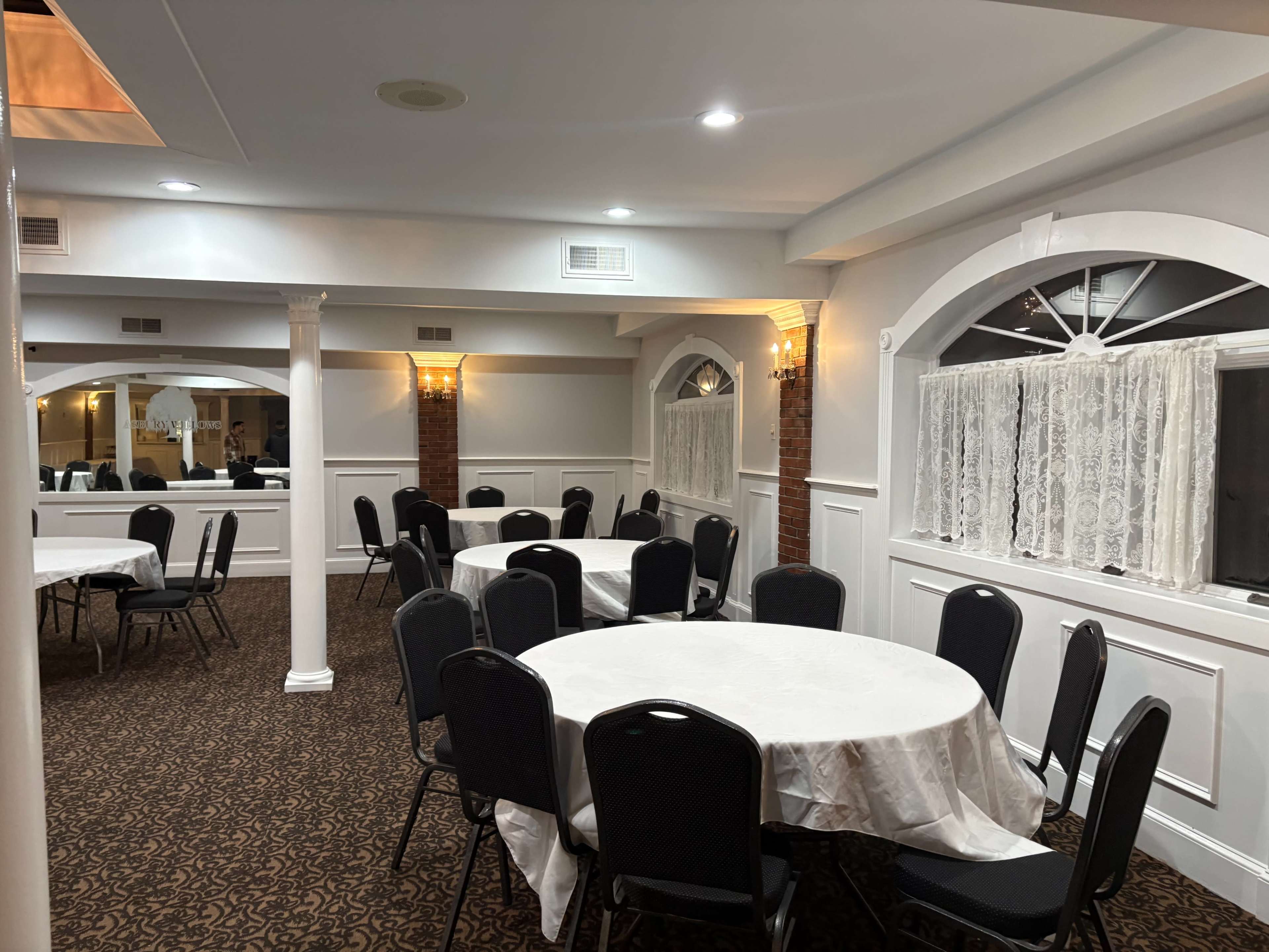 The image shows a well-lit dining room with several round tables covered in white tablecloths and black chairs arranged throughout the space.