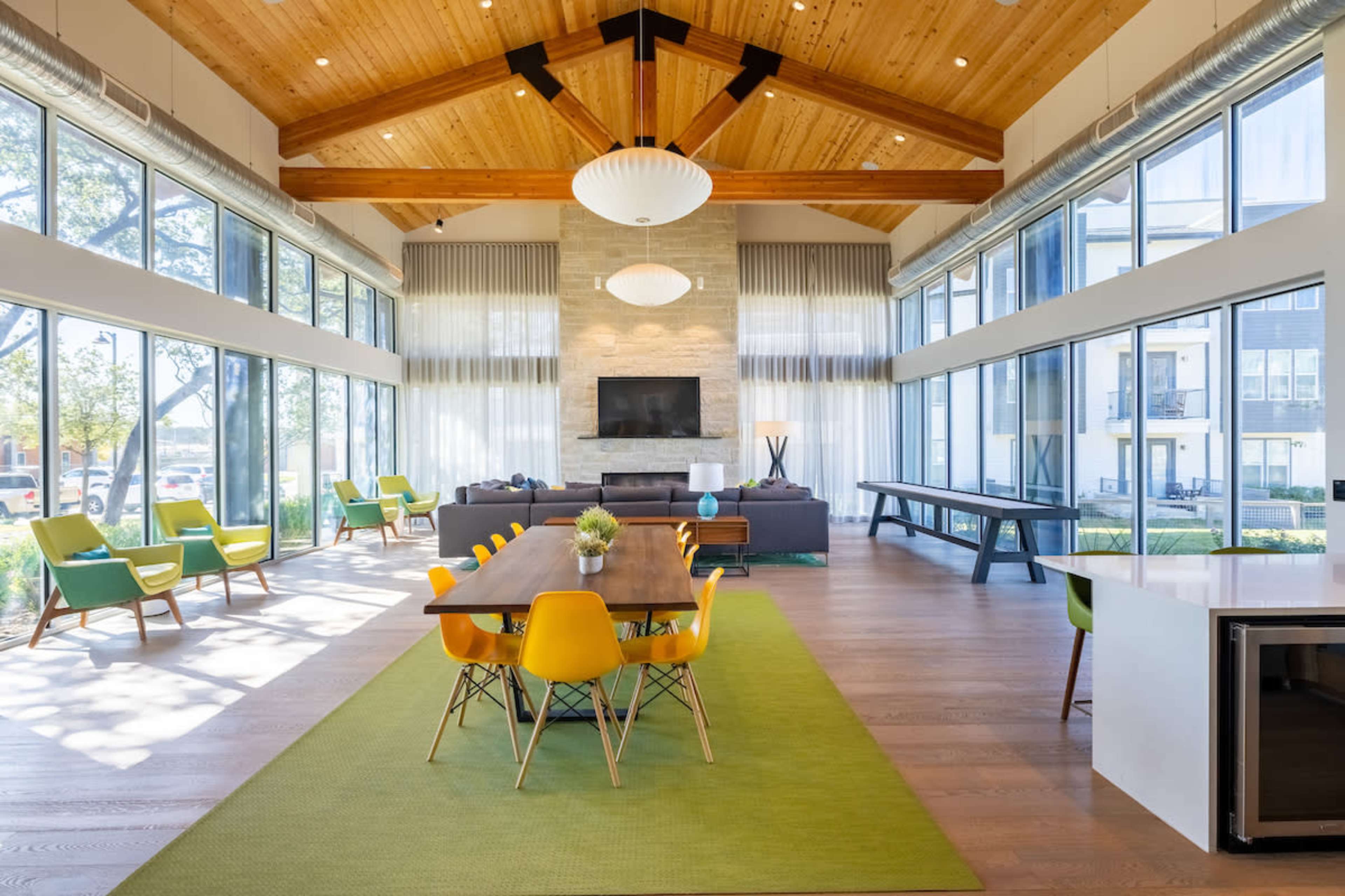 A spacious, modern lounge area with large windows, hardwood floors, a wooden ceiling, and a long dining table surrounded by colorful chairs.