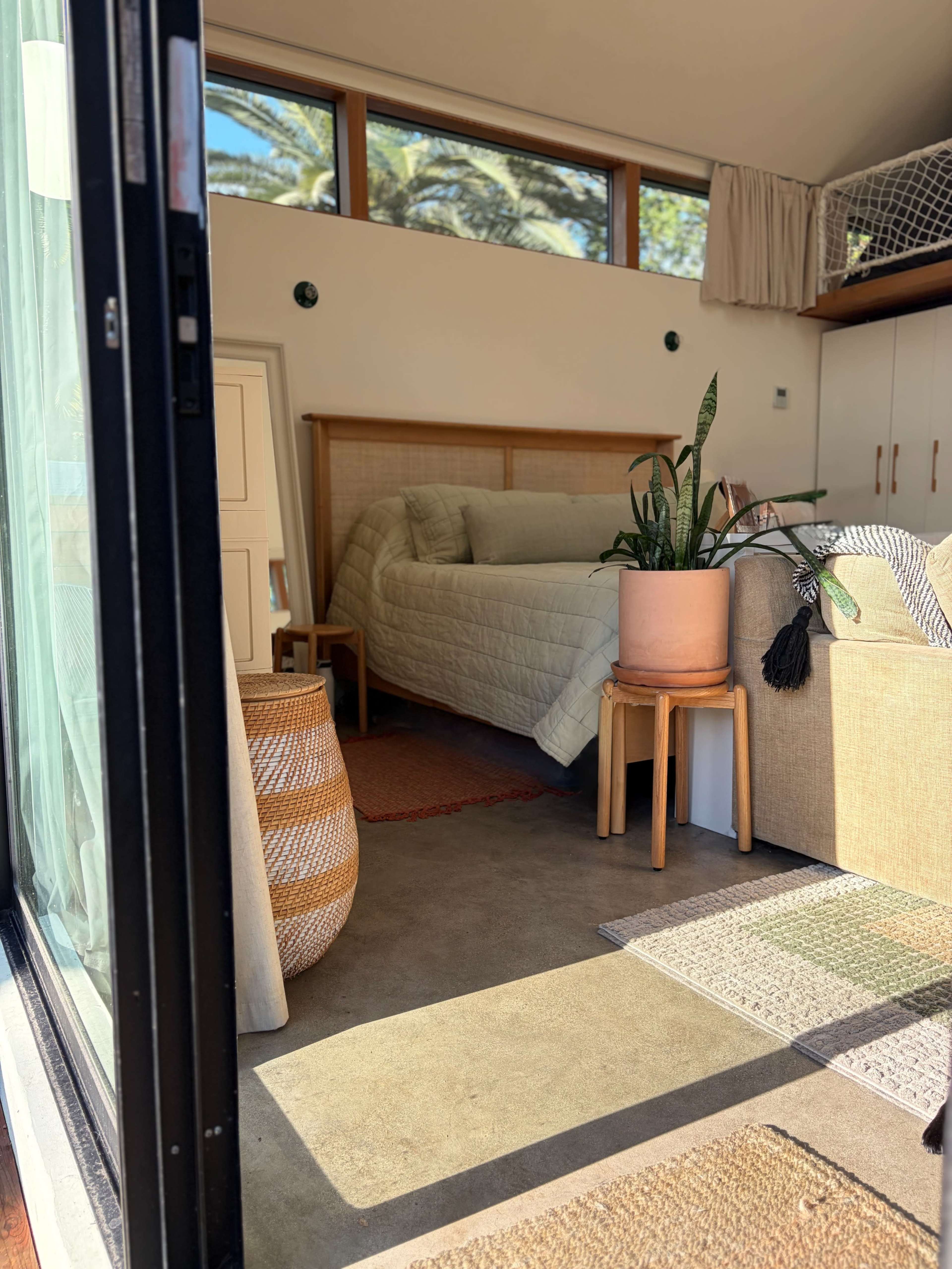 The image shows a cozy interior space with a bed, a sofa, plants, and various decorative elements arranged in a warm layout.