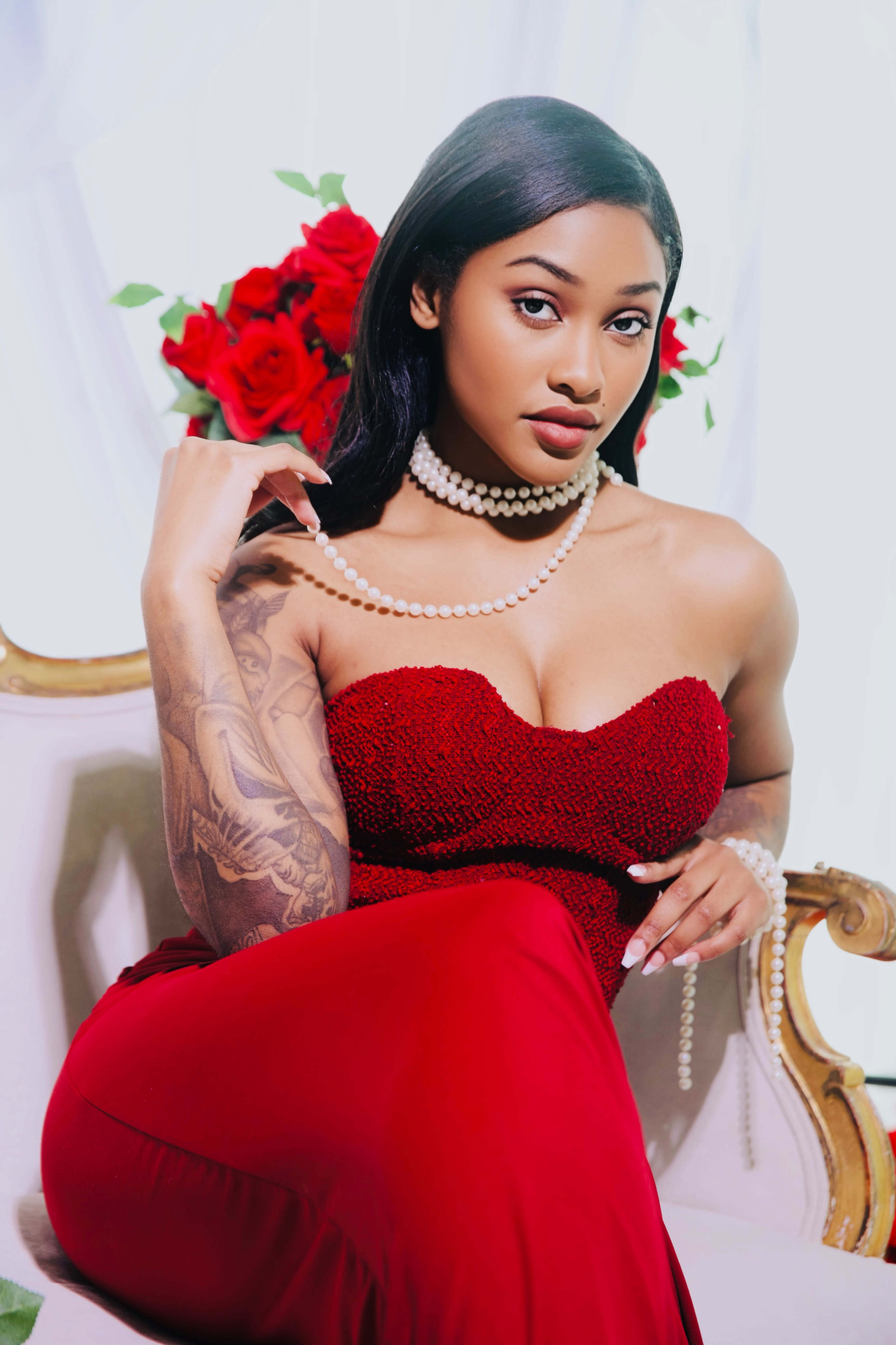 A woman in a red strapless dress poses on a chair, adorned with pearl necklaces and roses in the background.