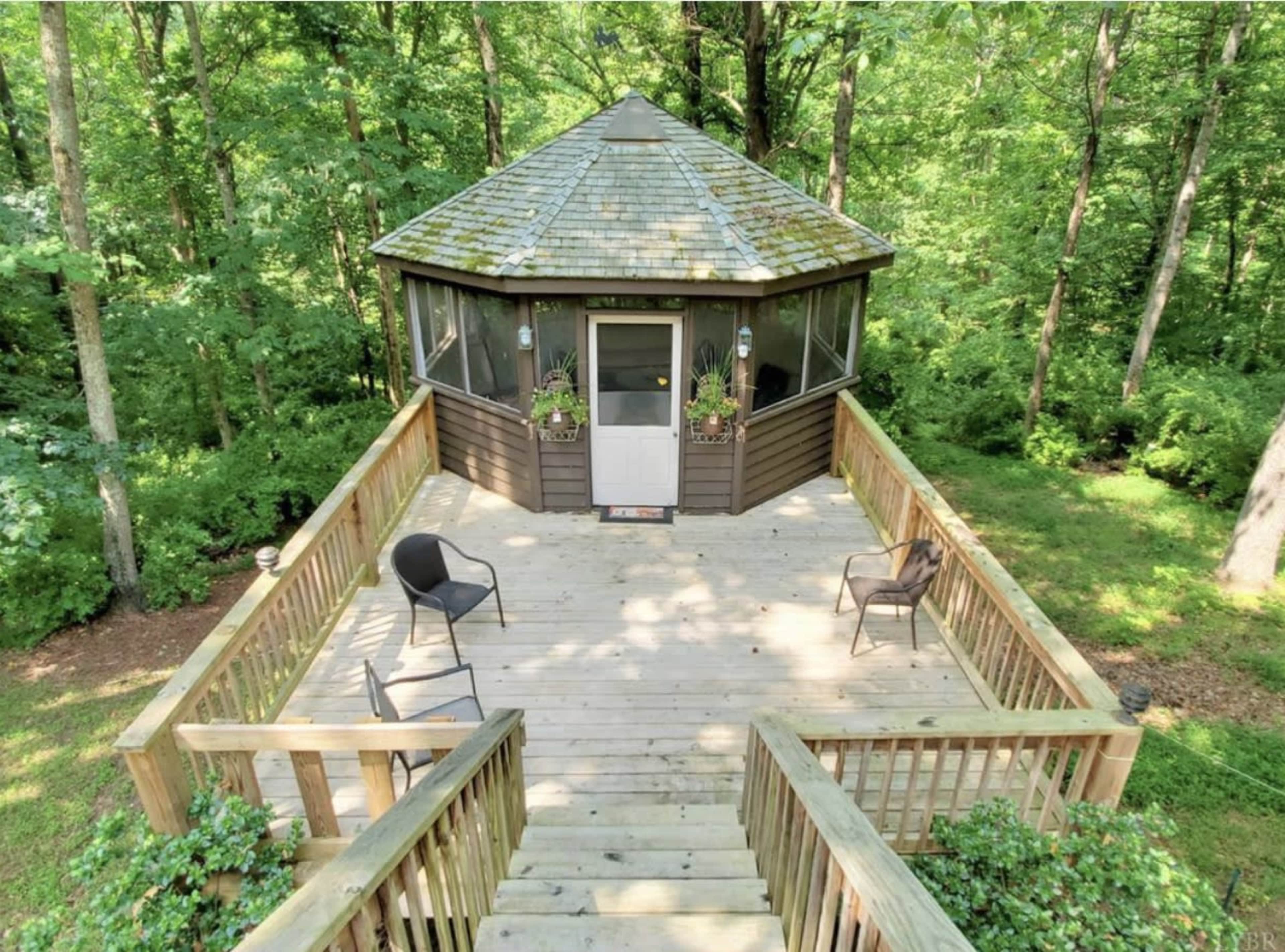A wooden deck surrounds a circular cabin with a shingled roof, situated in a forested area.