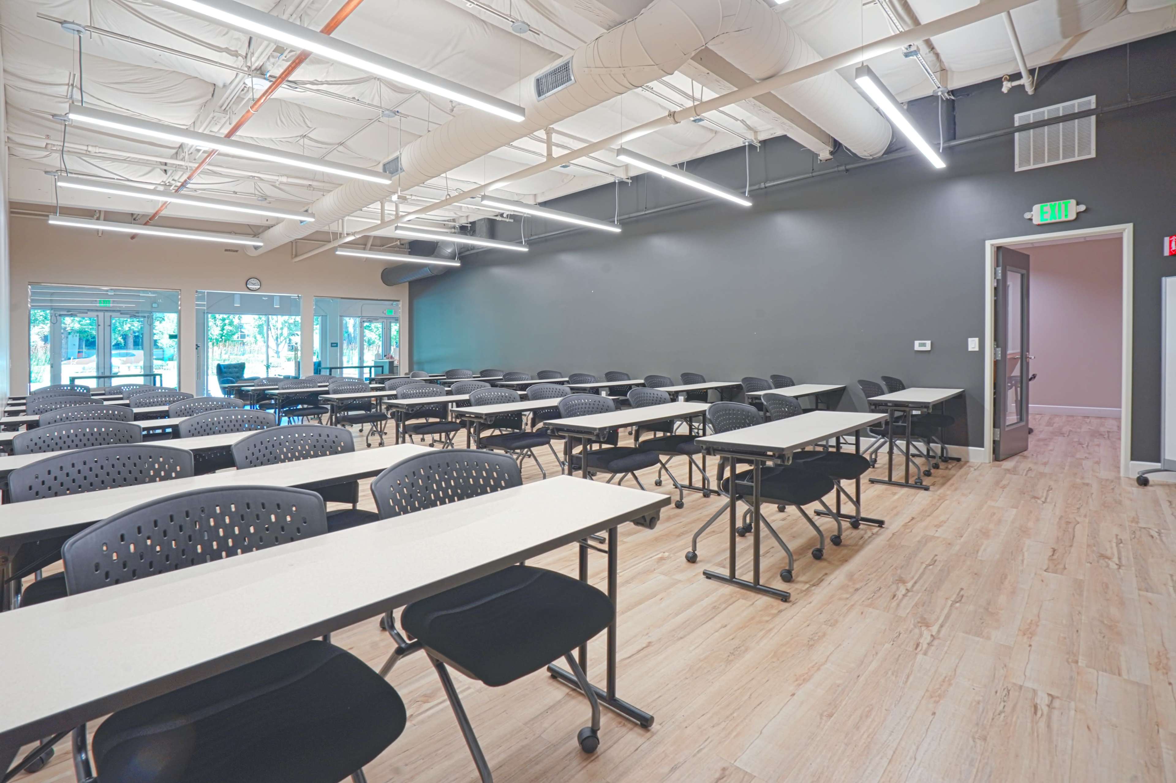 A modern classroom with multiple rows of black chairs and tables, featuring large windows and a door leading outside.