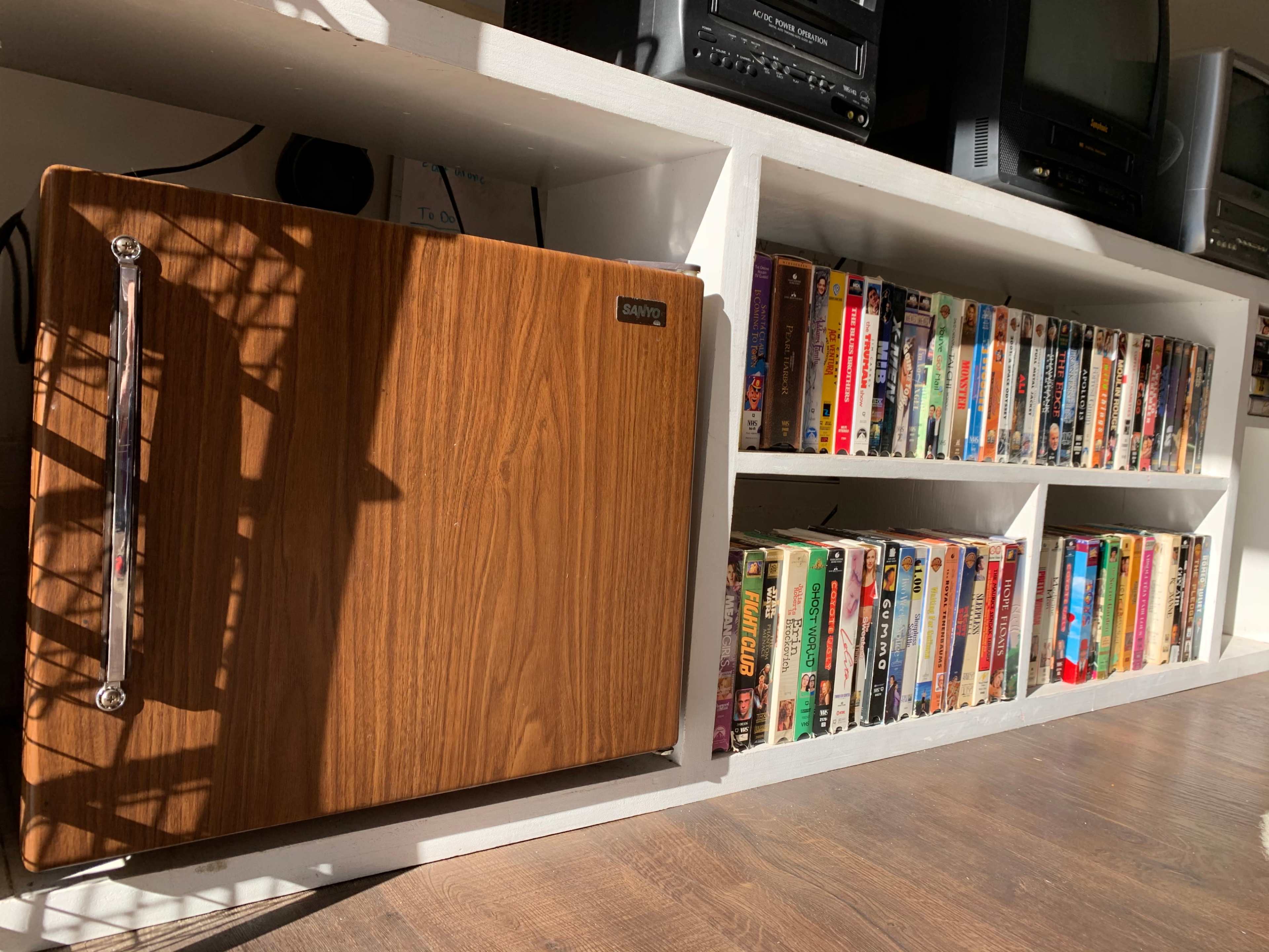 A wooden mini fridge placed beside a shelf filled with various VHS tapes.