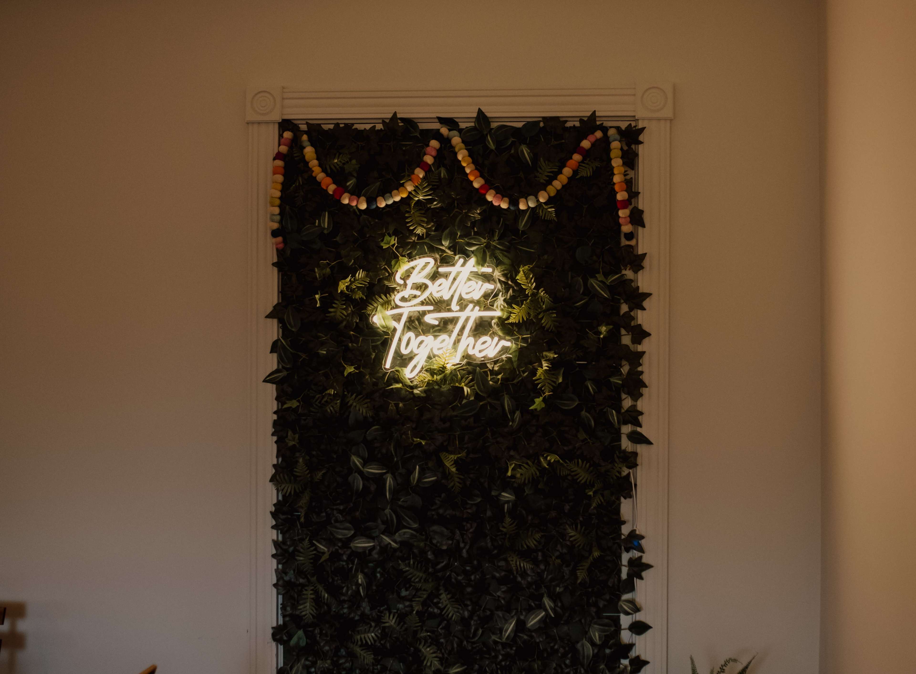 A neon sign reading "Better Together" is illuminated against a backdrop of green foliage and colorful garlands.