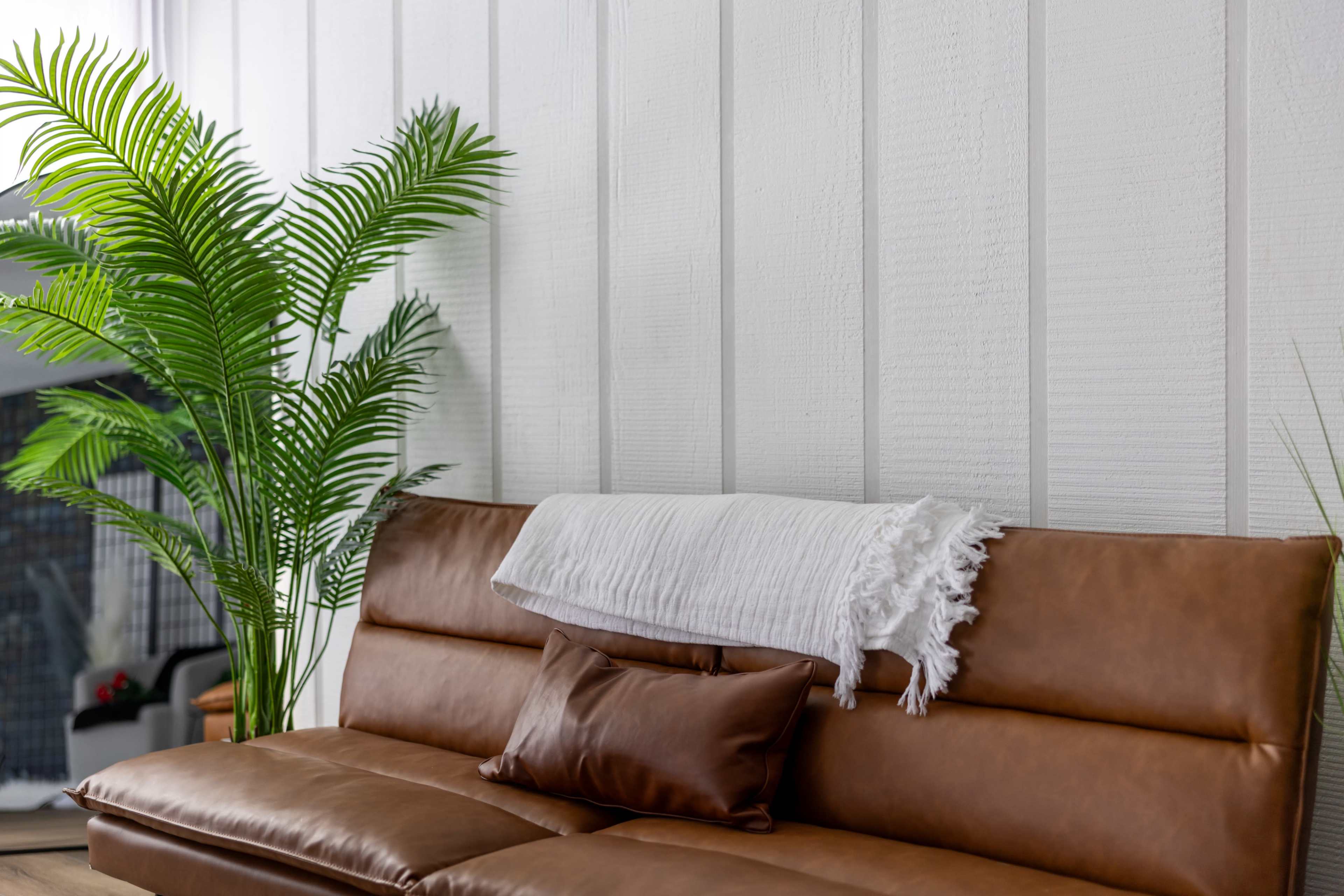 A brown leather sofa is positioned against a white-paneled wall, accompanied by a large potted fern and a white throw blanket draped over the backrest.