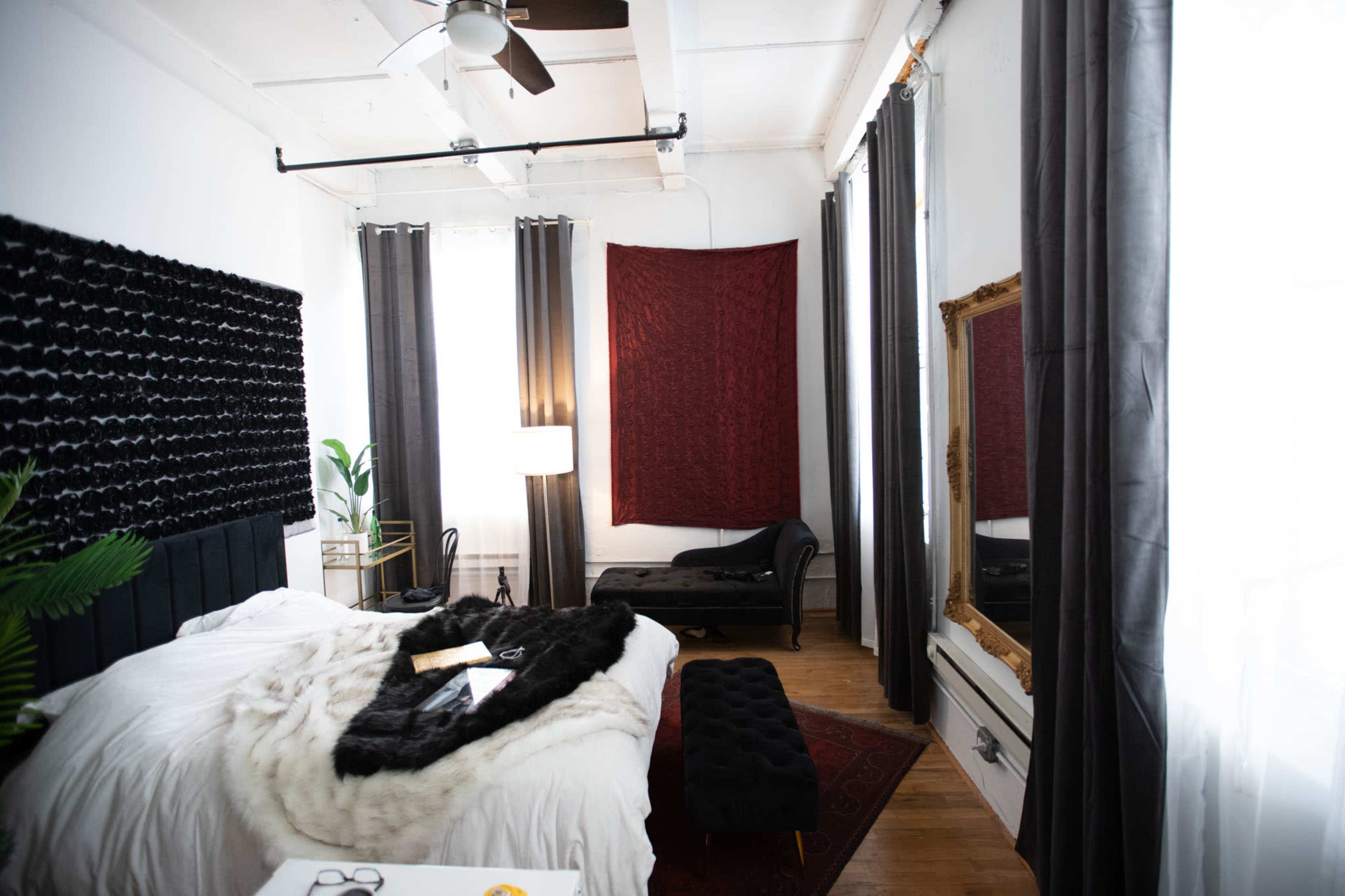 A modern bedroom with a large bed, a decorative black wall panel, a chaise lounge, and a mirror, all illuminated by natural light.