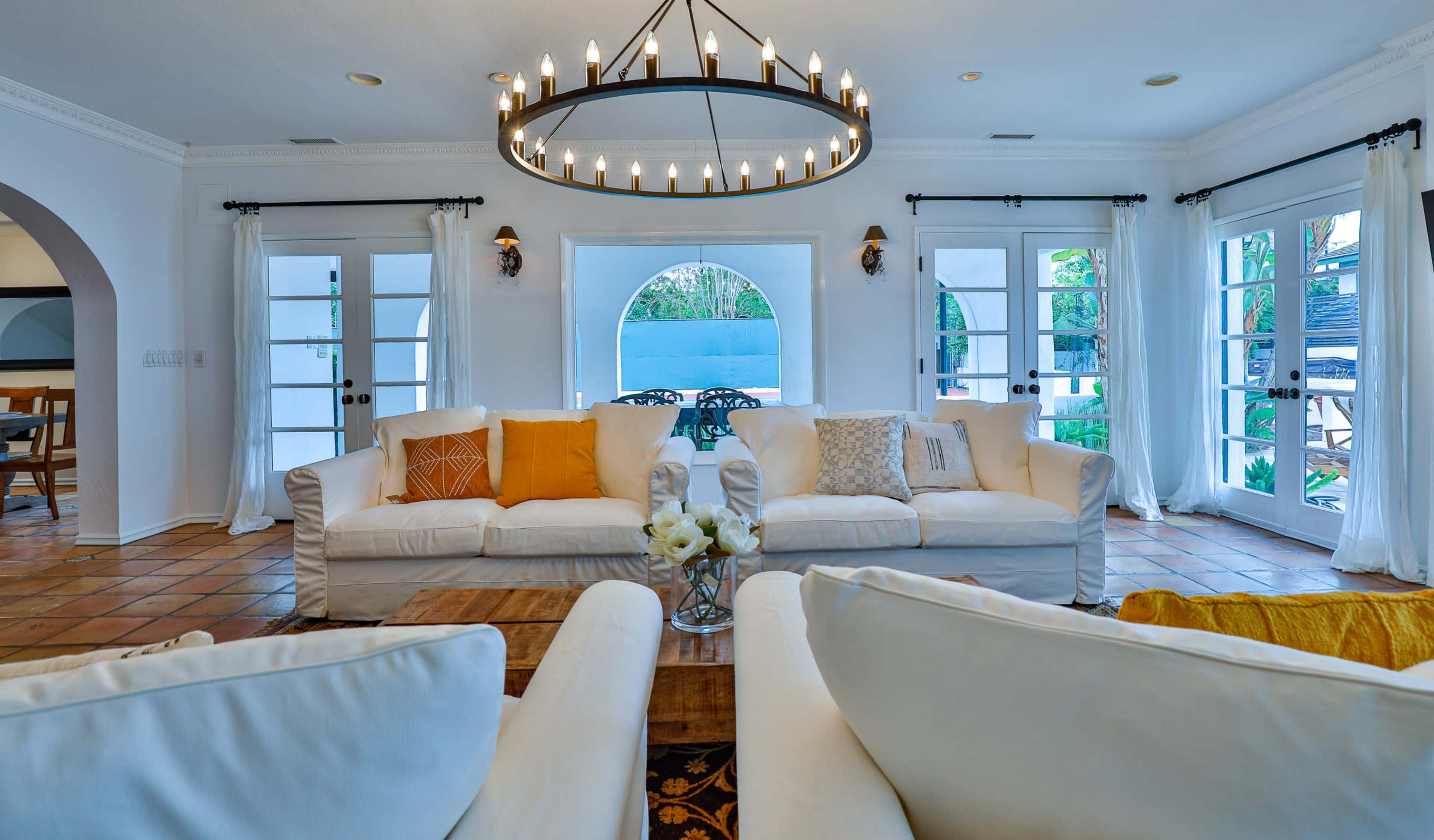 The image shows a spacious living area featuring white sofas, a large circular chandelier, and large glass doors leading to an outdoor space.