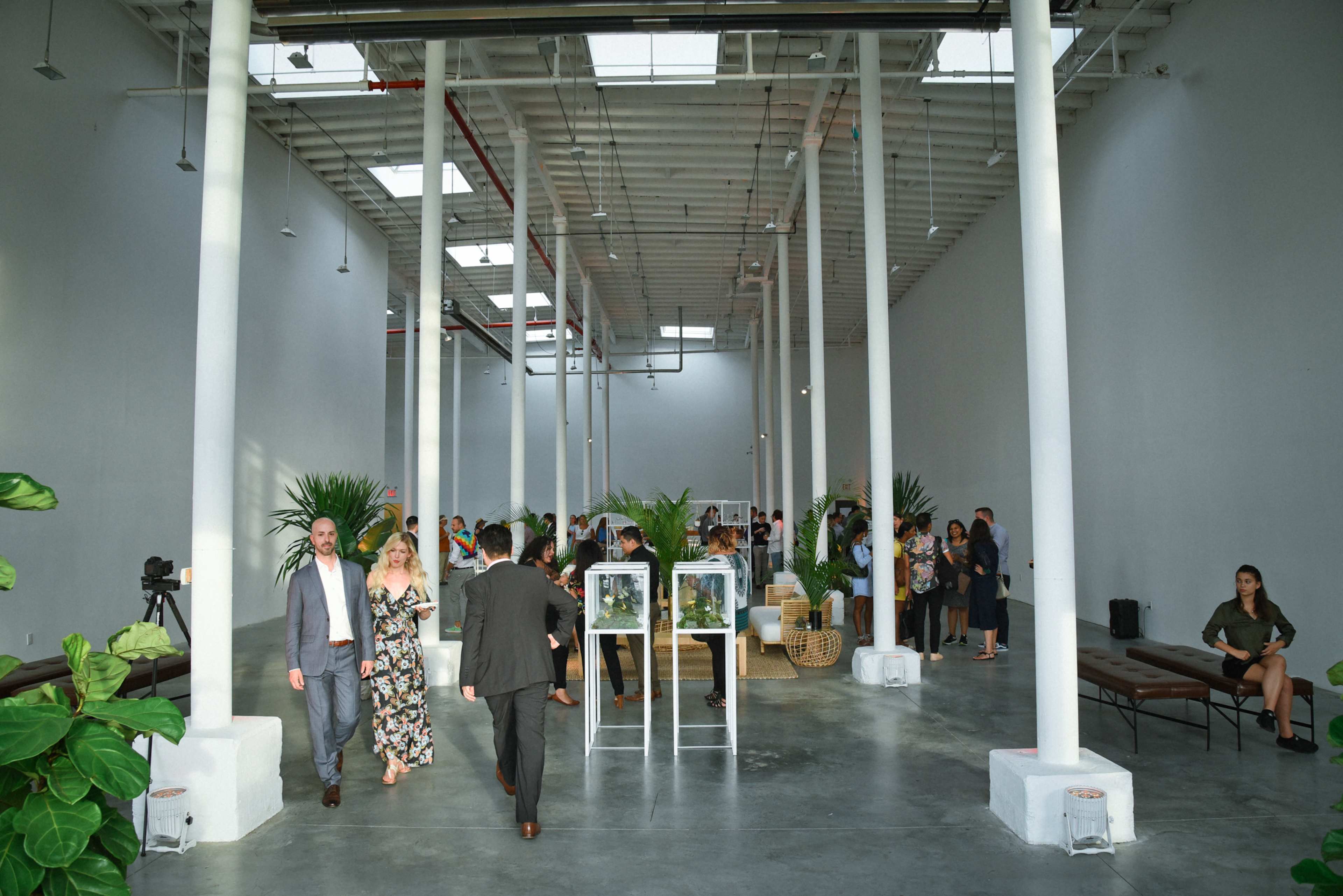 A spacious, brightly lit event venue features guests mingling, tall white columns, and modern seating arrangements.