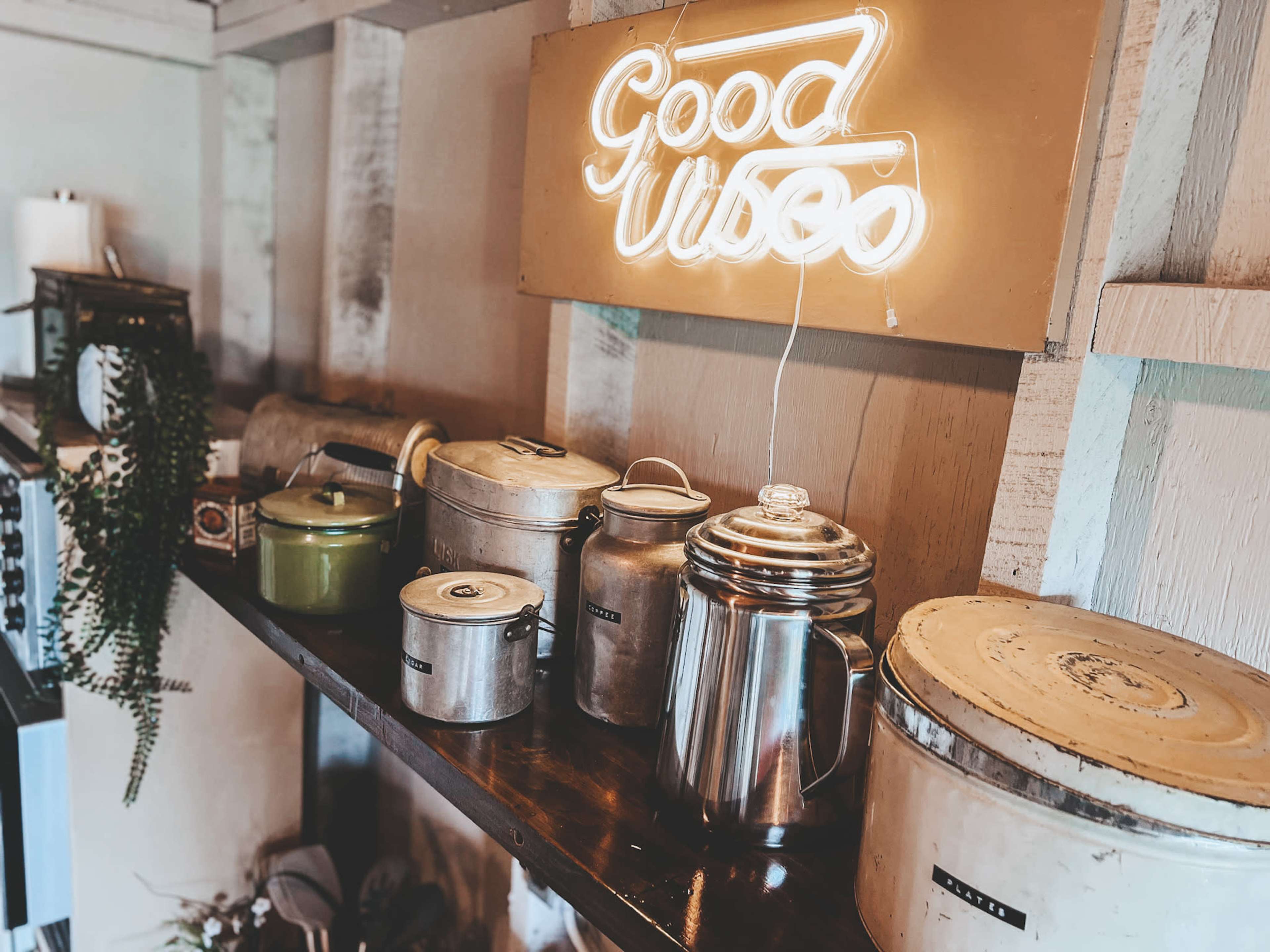 A wooden shelf displays various metal and ceramic containers, with a neon sign above that reads "Good Video."