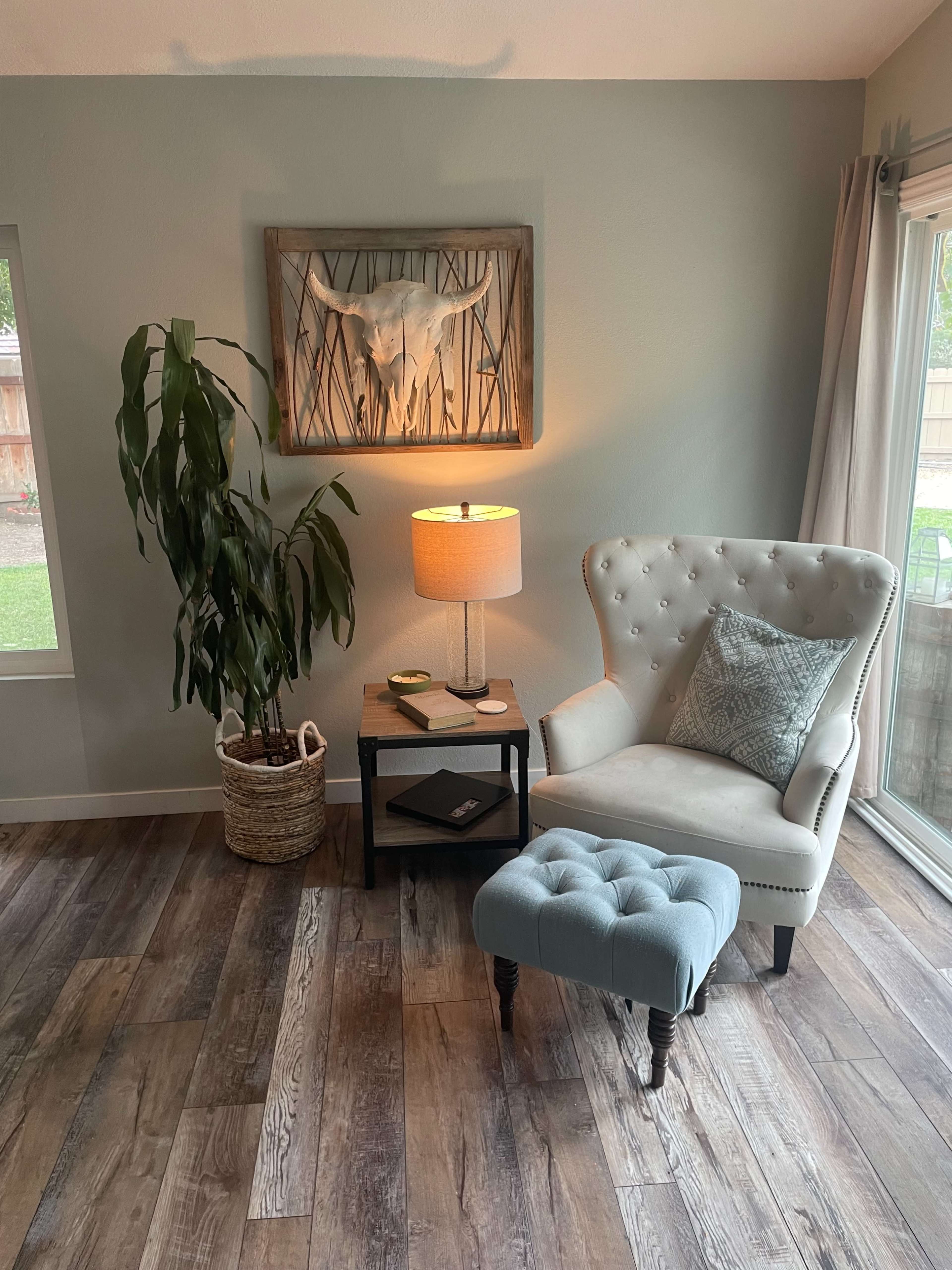 A cozy corner features a tufted armchair, a small side table with a lamp, and a potted plant beside a window, all set on wooden flooring.