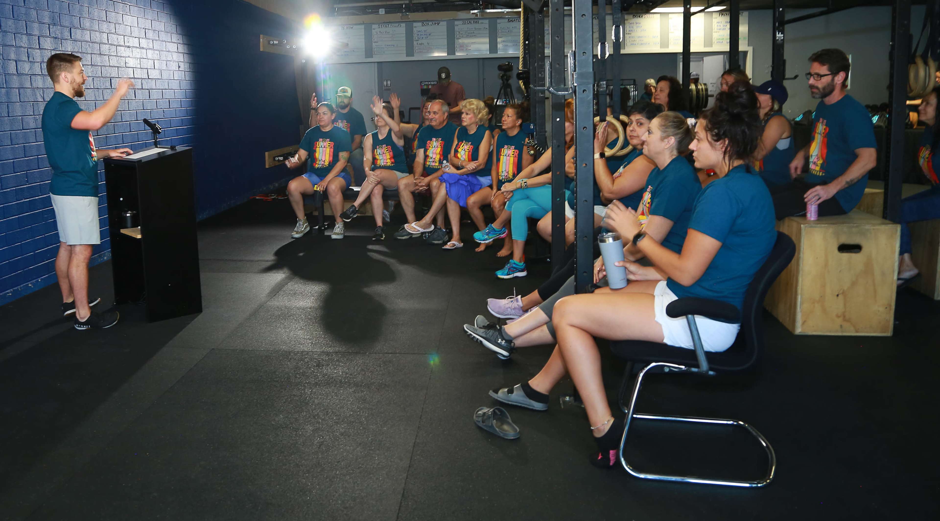 A speaker addresses an audience of people wearing matching shirts seated in a gym setting with exercise equipment in the background.