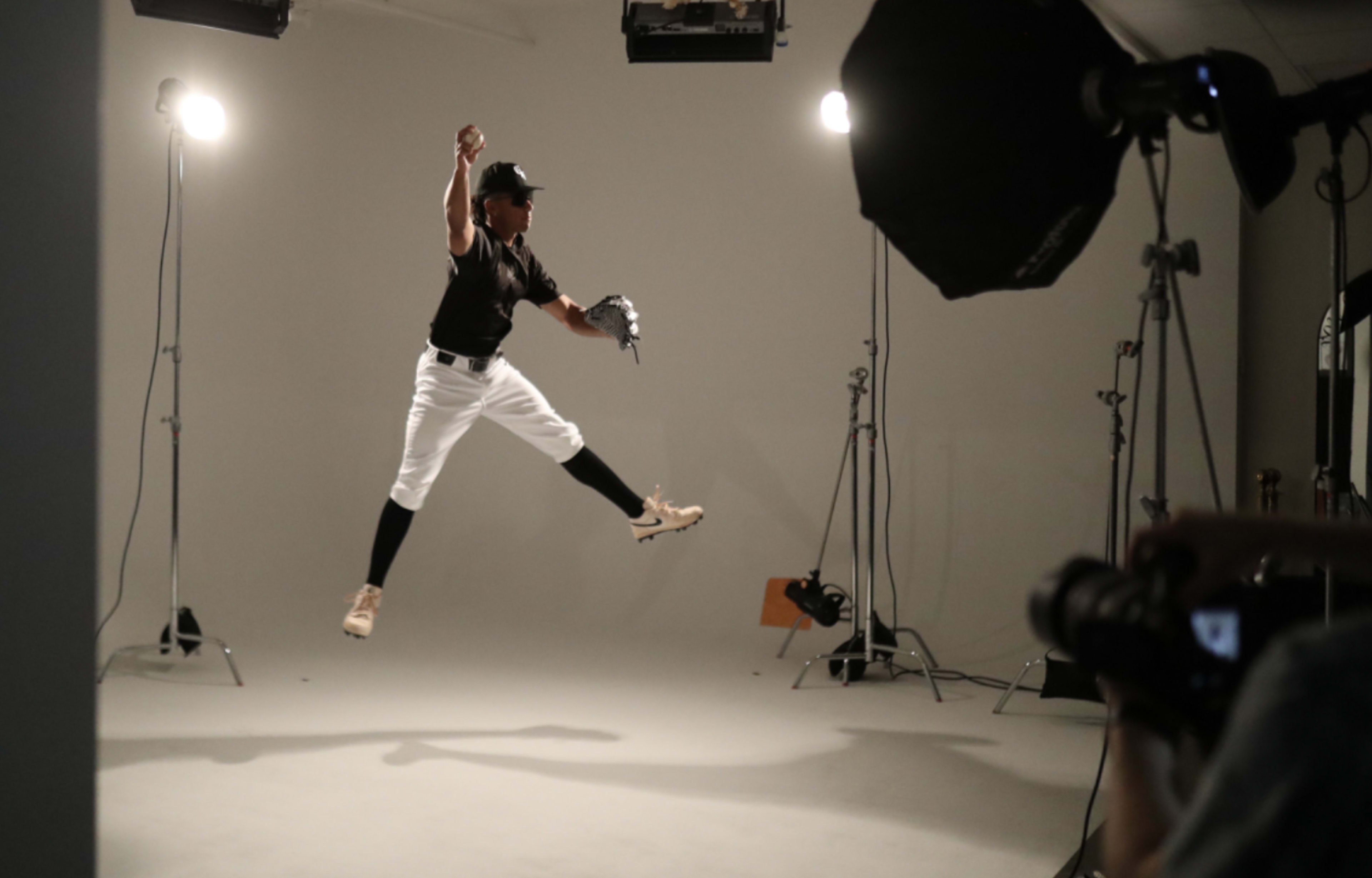 A baseball player in a black uniform jumps mid-air while preparing to throw a baseball in a studio setting.