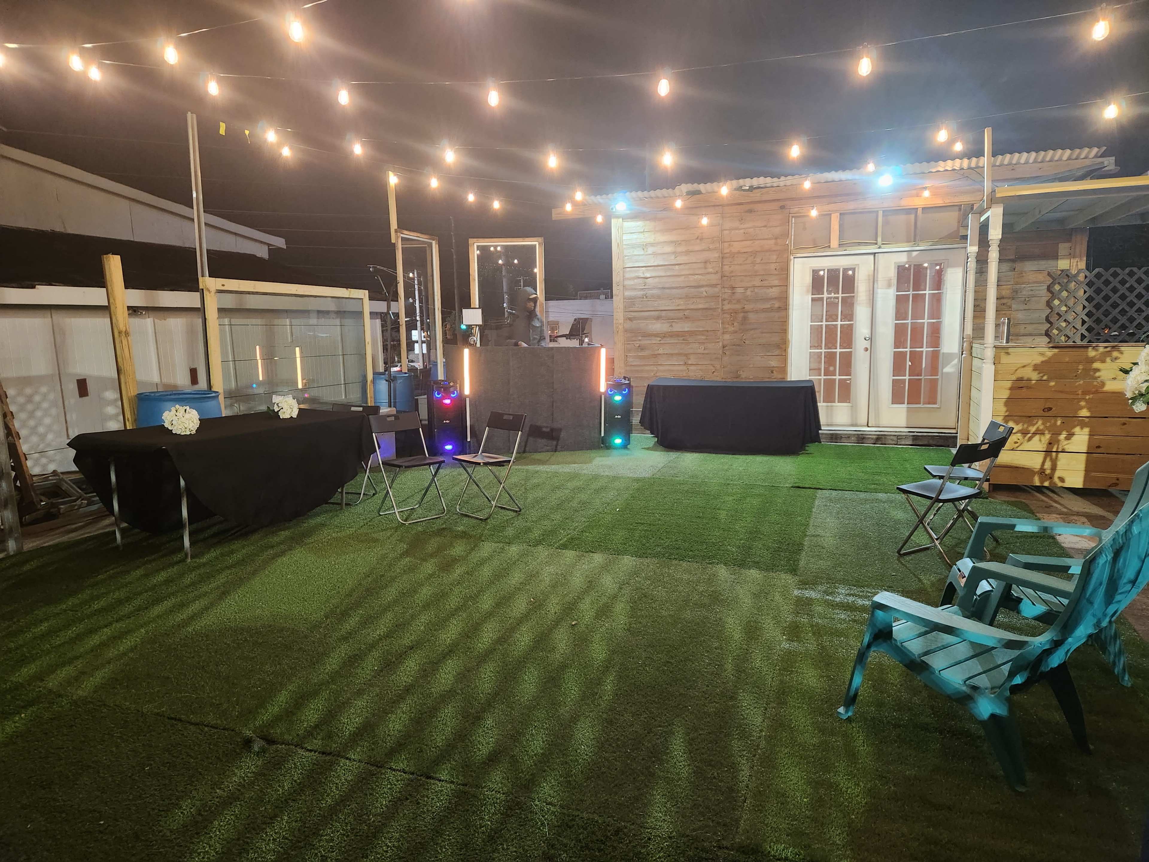 The scene shows a backyard event space at night, featuring string lights overhead, a bar area, and various chairs arranged on artificial grass.