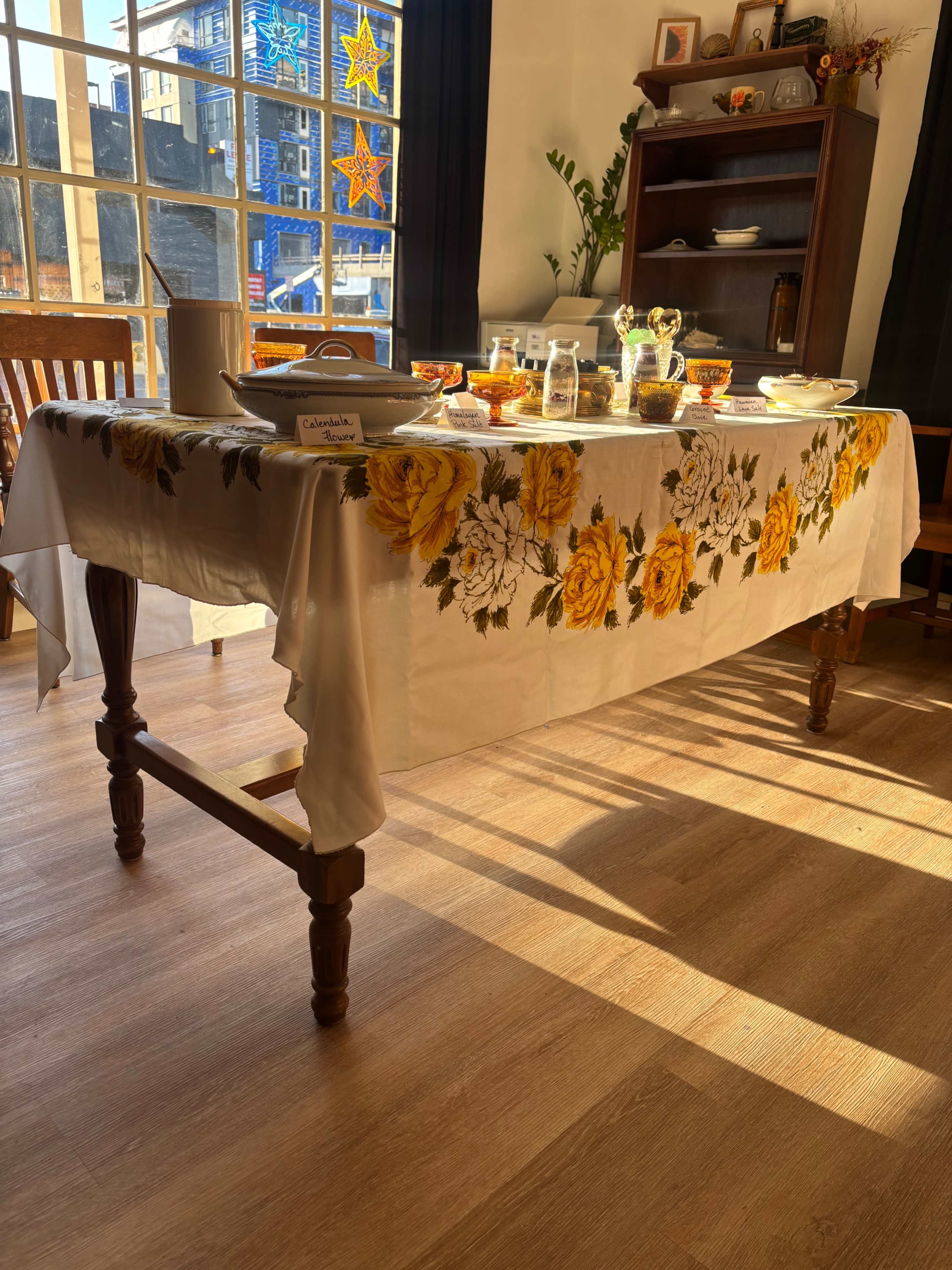 A dining table covered with a floral tablecloth is set with dishes and drinks in a brightly lit room.